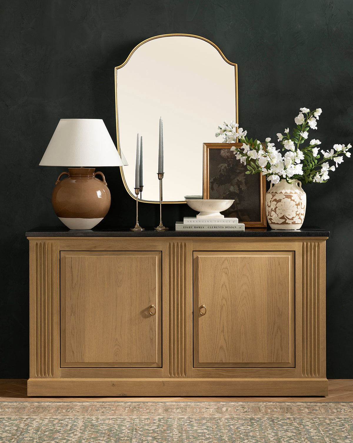 A wooden sideboard with two cabinets stands against a dark wall, topped with a lamp, candles, books, bowl, framed photo, and white flowers. Above it all hangs the elegant Sussex Wall Mirror by McGee & Co.