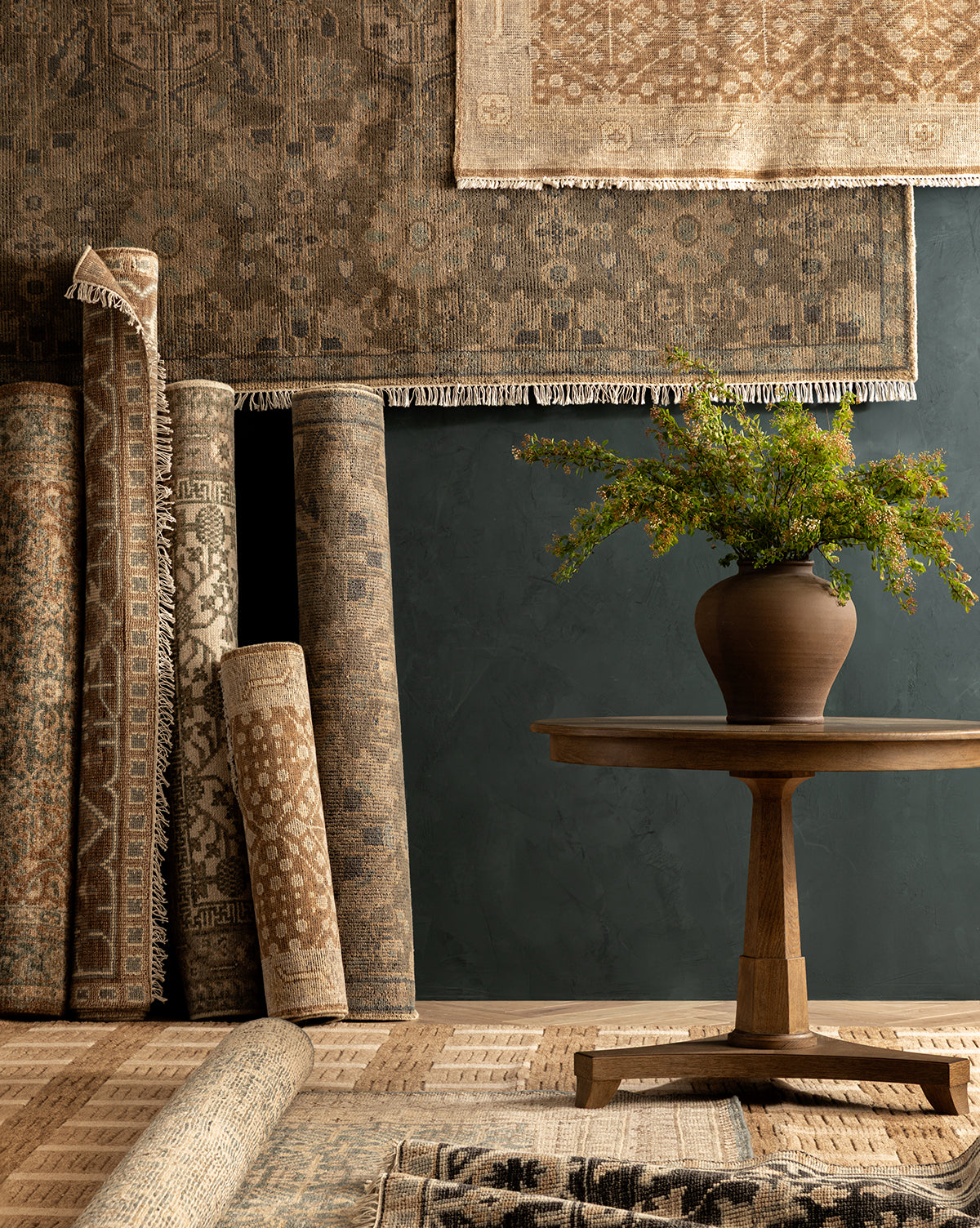 Several Endicott Hand-Tufted Wool Rugs in neutral patterns are rolled and hanging against a dark wall, with a wooden table holding a brown vase of green foliage placed in front, atop a textured rug floor by McGee & Co.
