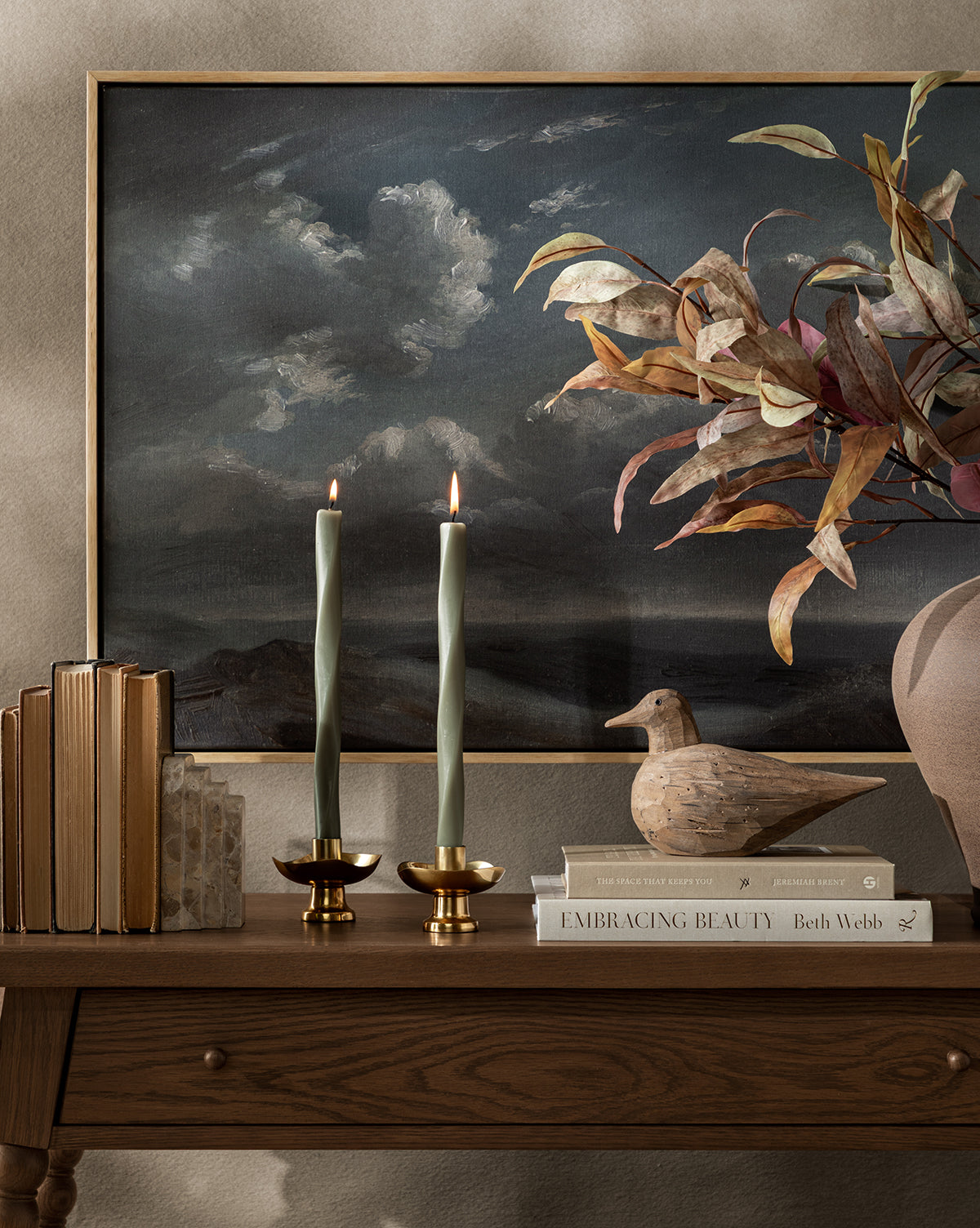 A wooden table holds two lit brass candle holders, a stack of books, a wooden bird figurine, and a large vase filled with Faux Purple Eucalyptus Stem, set against a backdrop of a cloudy sky painting, by McGee & Co.