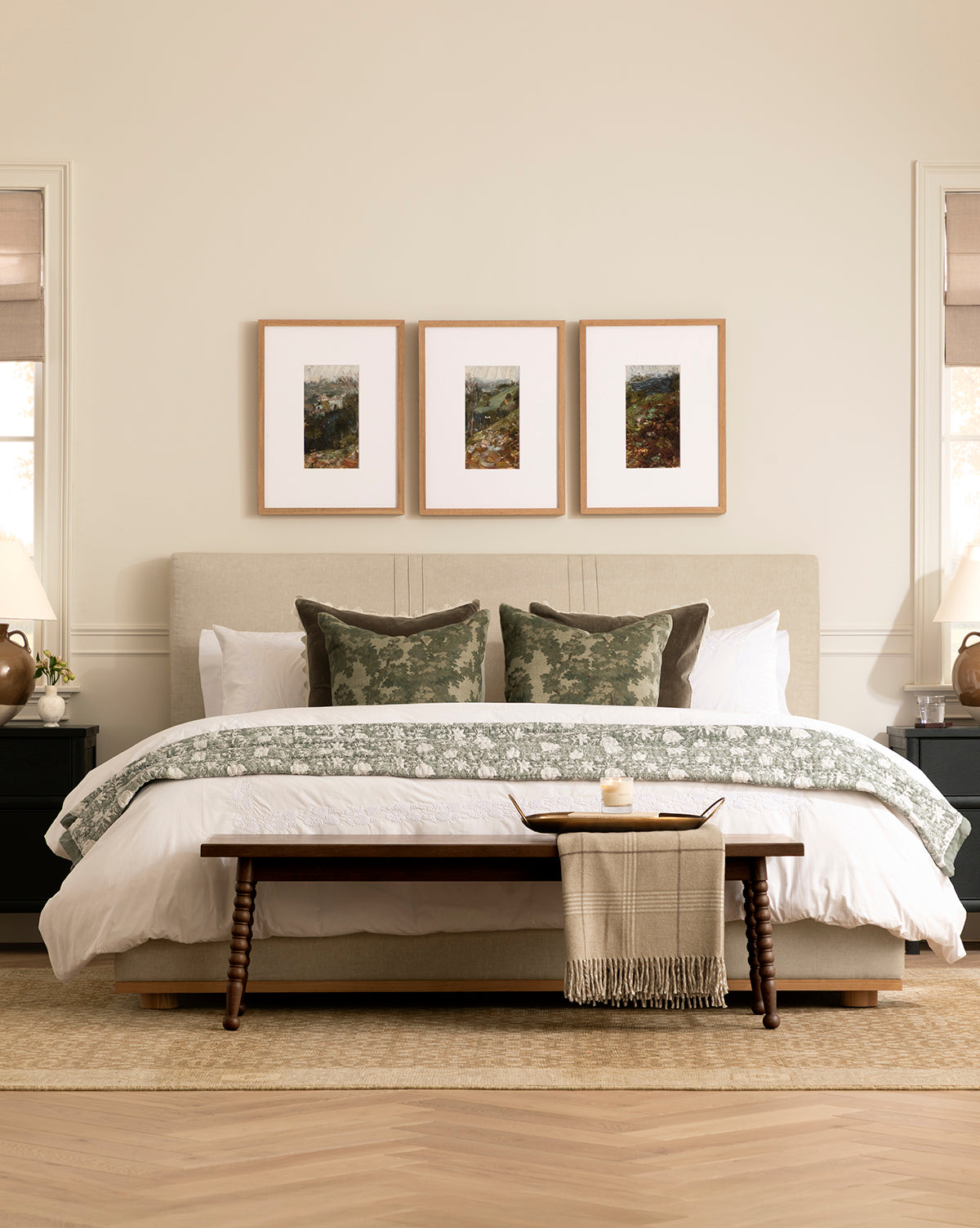 A neatly made bed with green and white pillows and a Nottingham Throw stands in a cozy bedroom. Above the bed are three framed artworks, with nightstands, lamps, décor on each side, and a bench placed at the foot, by McGee & Co.