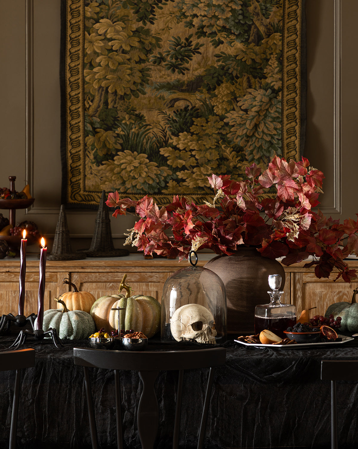 A dark, elegant table is decorated for autumn with pumpkins, a skull under a glass dome, candles, treats, and a Faux Autumn Grapevine Stem in a vase. An ornate tapestry with foliage hangs on the wall behind this seasonal arrangement, by McGee & Co.