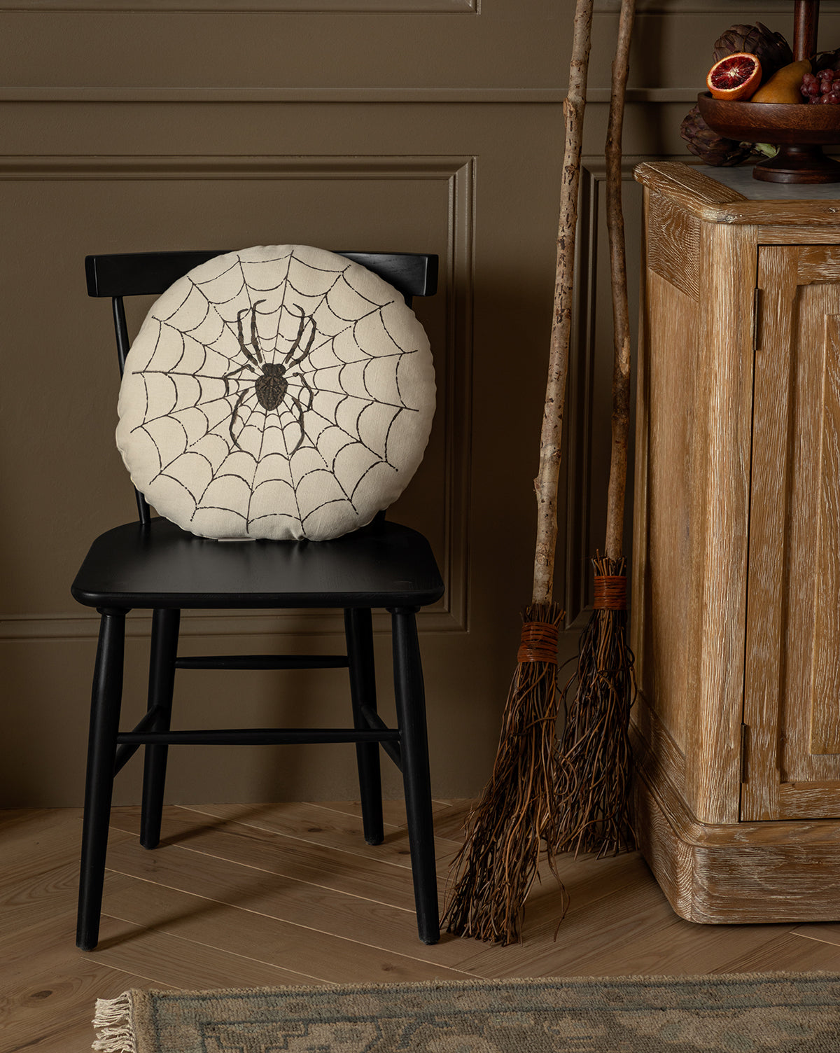 The Spider Pillow, featuring an embroidered spider design, rests on a black wooden chair while two rustic brooms lean against a cabinet—creating a cozy autumn or Halloween accent ideal for seasonal décor, by McGee & Co.