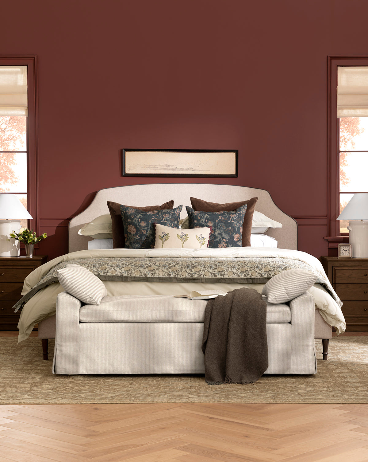 A cozy bedroom featuring a Willoughby Bed with linen upholstery, decorative pillows, and an English Oak bench at the foot. Wooden nightstands with lamps flank the bed, art hangs above, and deep red walls complete the inviting space, by McGee & Co.