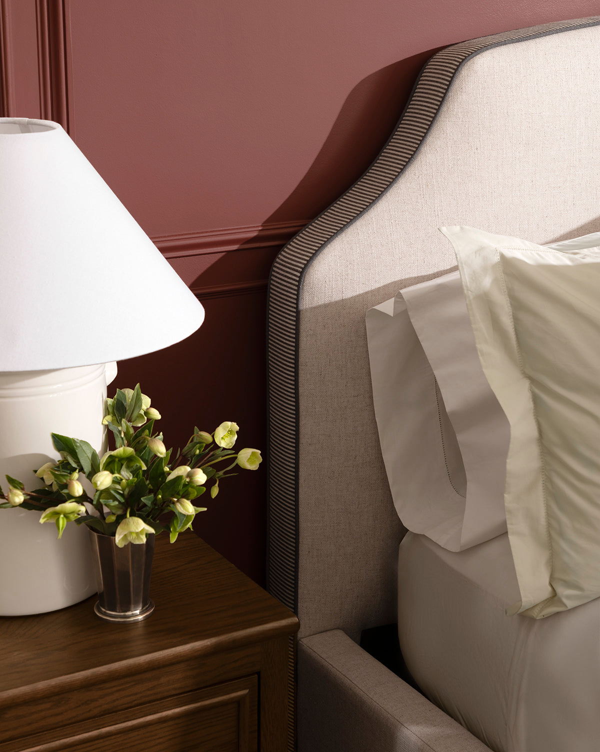 A close-up of a bedroom showcases the Willoughby Bed with linen upholstery, accented by white and light green pillows, a wooden nightstand, a white lamp, and a small flower vase against a mauve wall, by McGee & Co.
