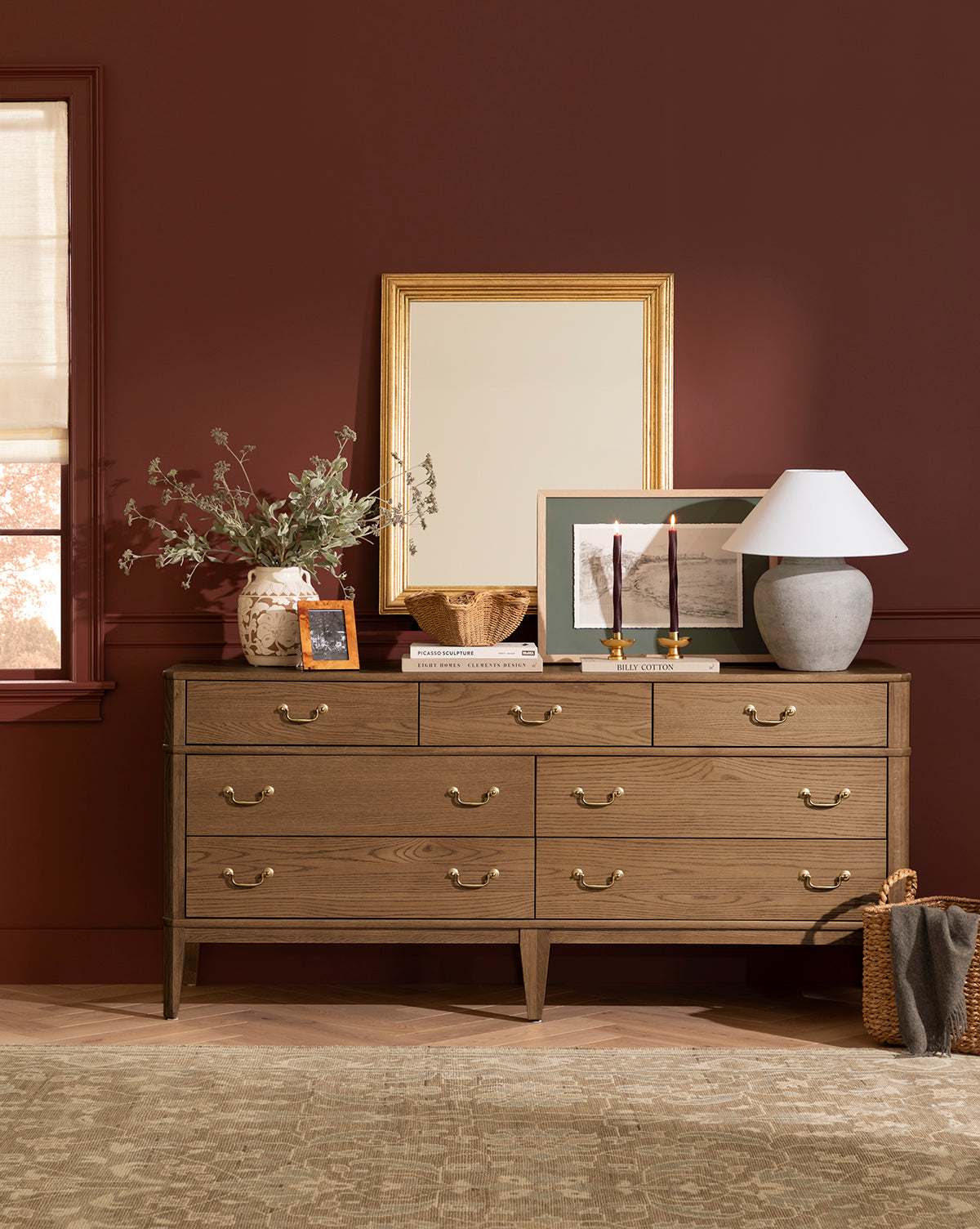 A dresser with brass handles stands against a burgundy wall, topped with the Suzanne Wall Mirror, a lamp, candles, framed art, a vase of greenery, a bowl, and a photo. A woven basket is placed on the floor next to the dresser by McGee & Co.