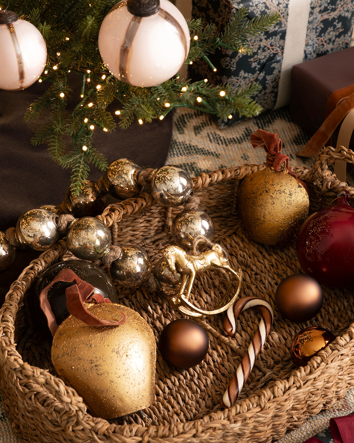 A woven basket holding gold and red Christmas ornaments, a Gold Rocking Horse Ornament, a candy cane, and metallic garland sits under a decorated tree with gifts, adding festive charm to your holiday décor, by McGee & Co.