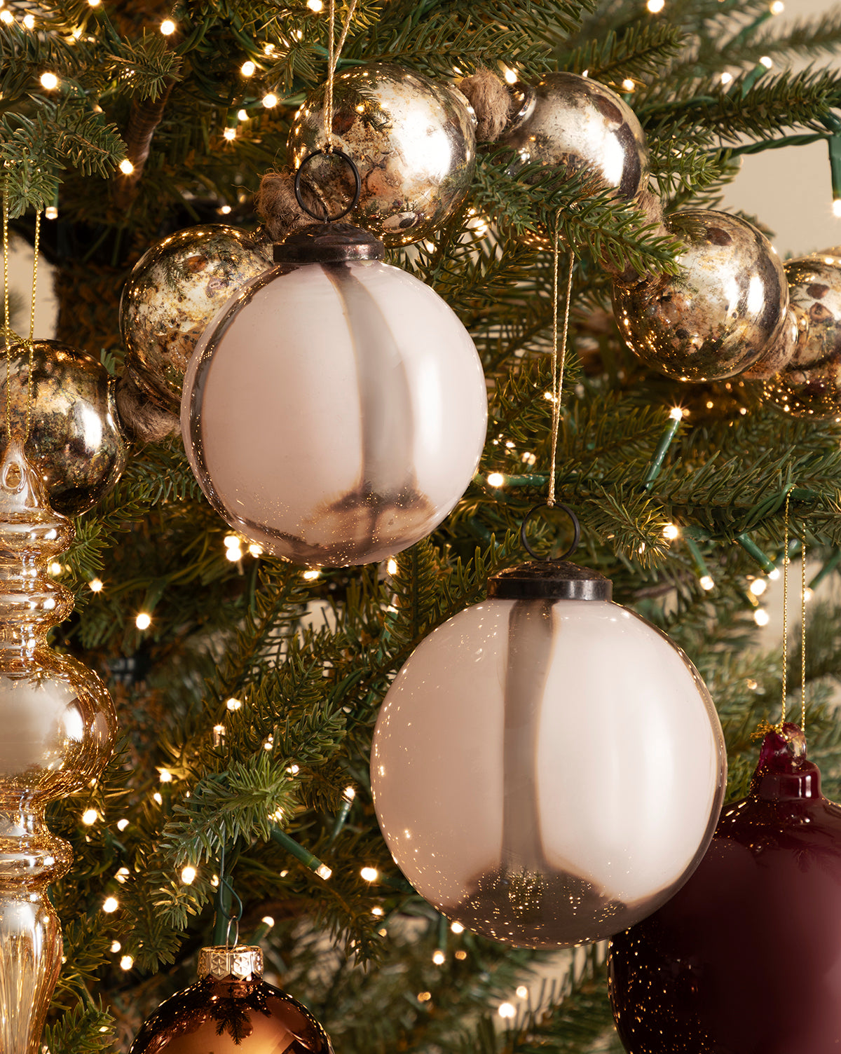 A 4" Striped Glass Bauble hangs on the holiday tree, aglow with white string lights and surrounded by gold and burgundy ornaments amid green pine needles, by McGee & Co.