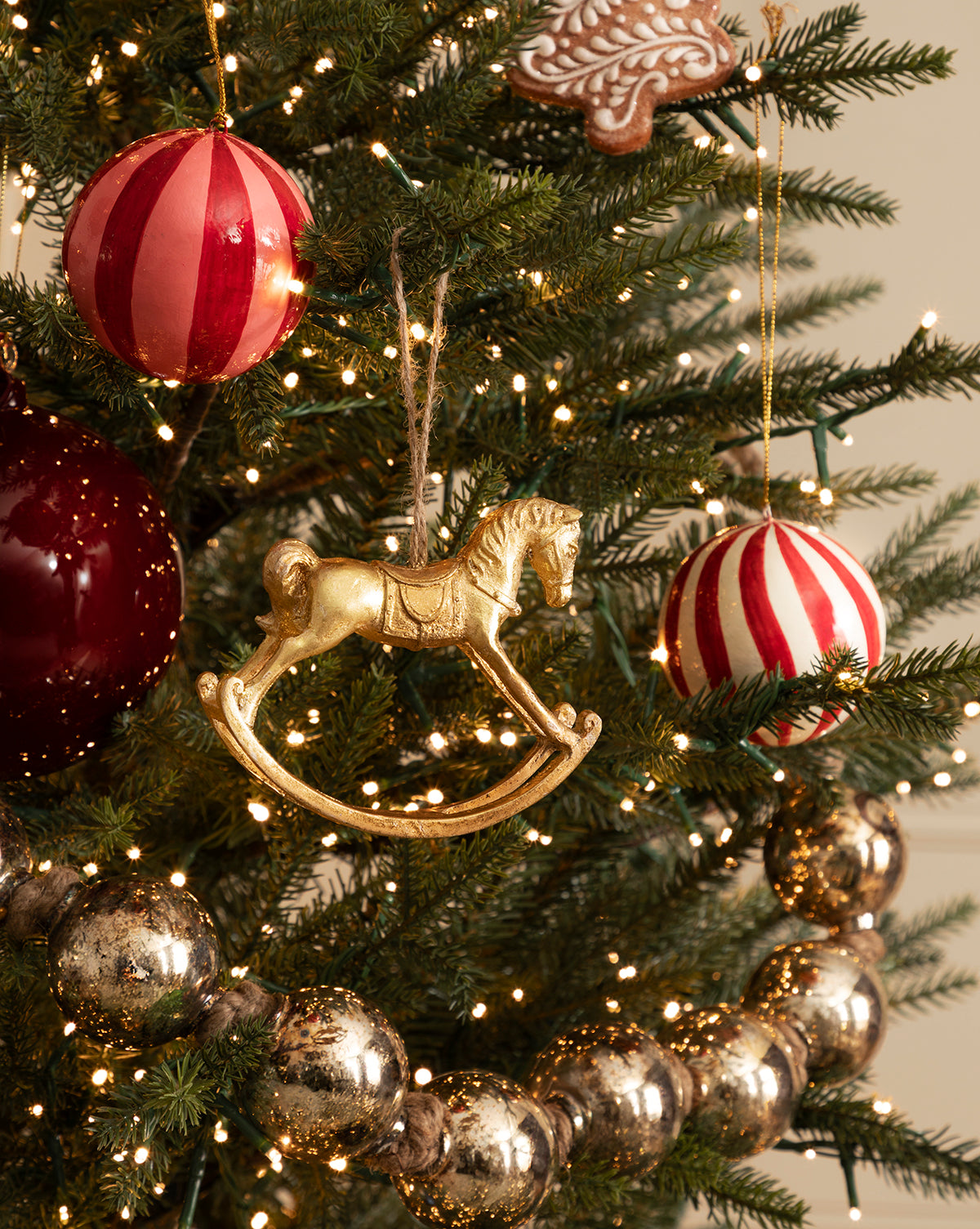A Christmas tree adorned with the Gold Rocking Horse Ornament, red and white striped ornaments, vintage baubles, metallic garland, and twinkling string lights brings festive charm to your holiday décor, by McGee & Co.