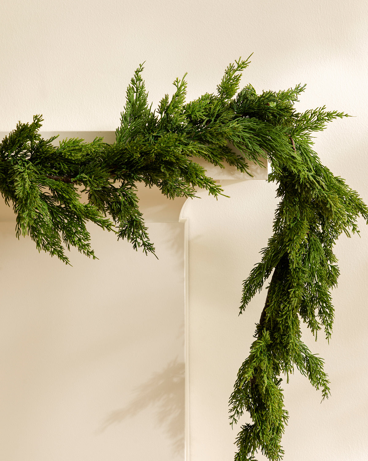 A 5' Cedar Faux Garland is draped over the corner of a white mantel against a neutral wall, creating the look of a lush, realistic garland, by McGee & Co.