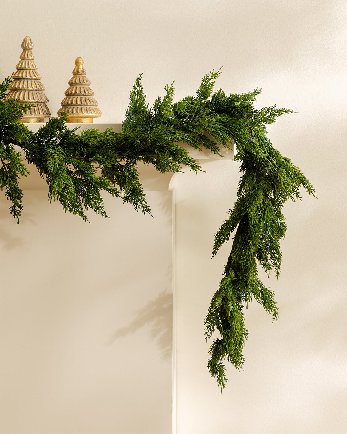 A 5' Cedar Faux Garland drapes over the edge of a white mantel, with two gold decorative Christmas tree figurines placed on top, by McGee & Co.