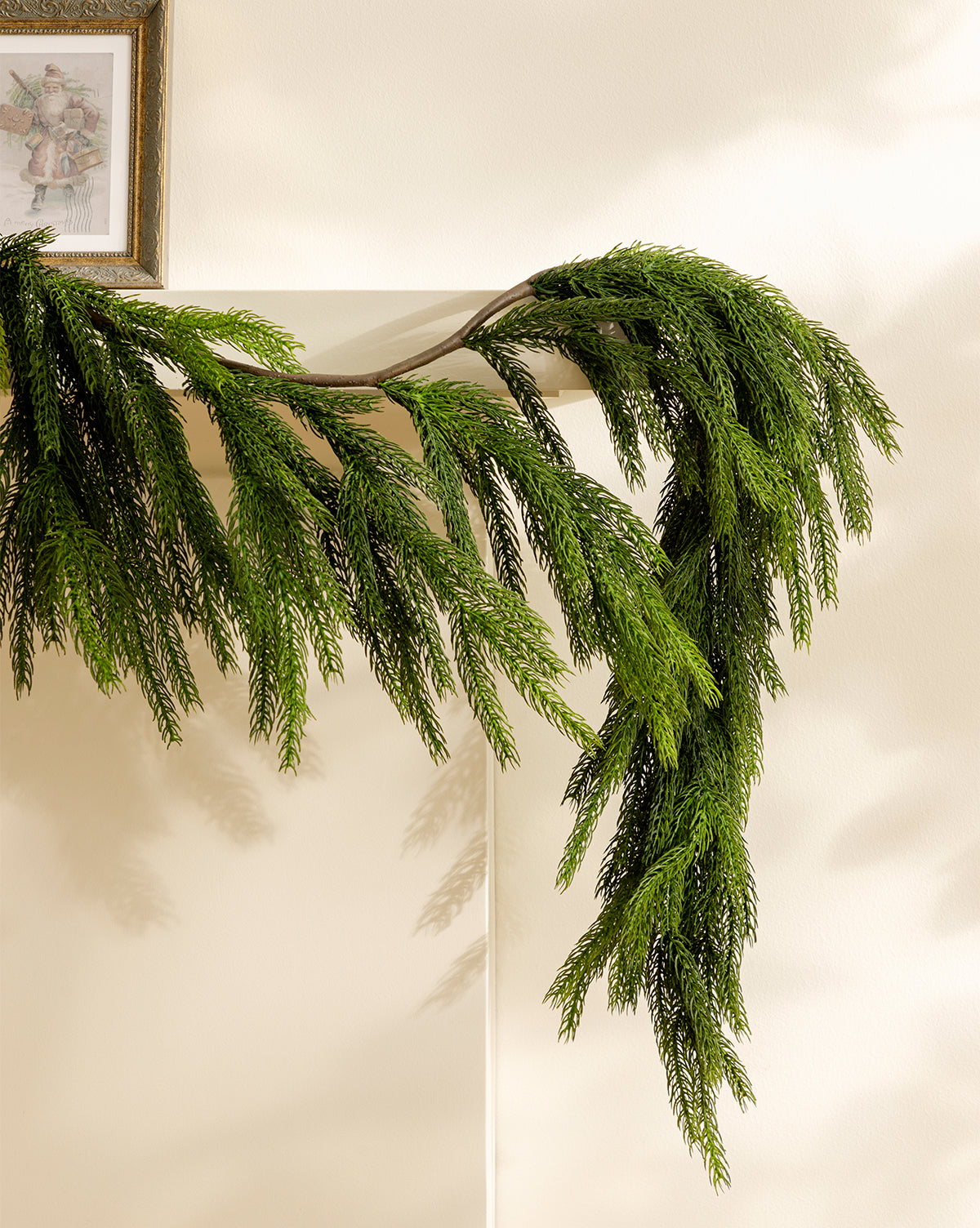 The 6' Norfolk Pine Faux Garland drapes over a light mantel, its realistic green branches casting shadows on the wall, while a framed floral picture hangs above the festive greenery, by McGee & Co.
