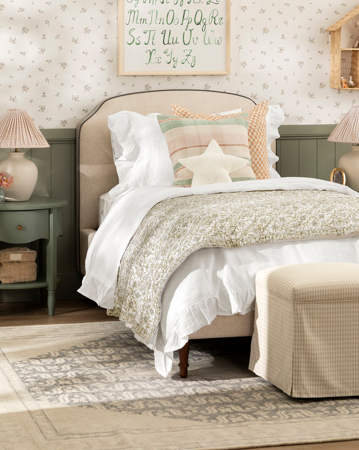 A cozy bedroom with a neatly made Willoughby Bed, white and pastel bedding, decorative pillows including a star-shaped one, English Oak legs, green nightstands with lamps, floral wallpaper, and an alphabet print above the headboard, by McGee & Co.