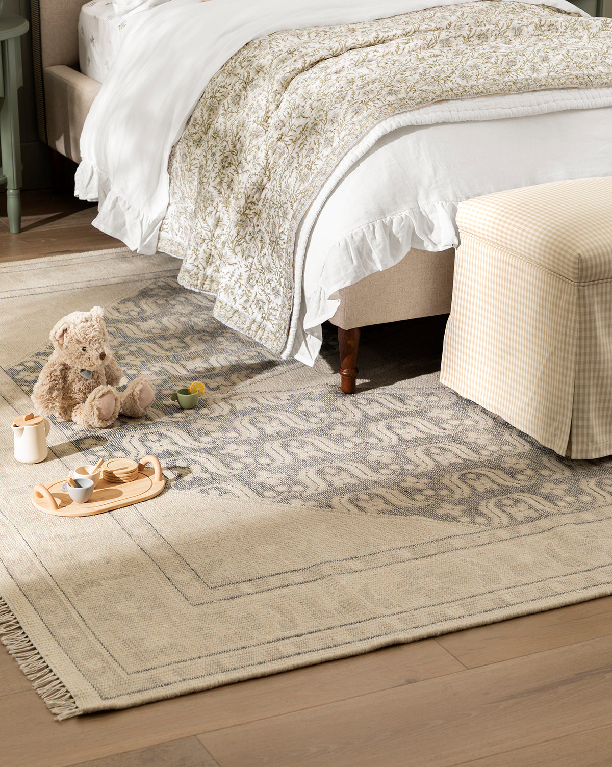 A cozy bedroom features a bed with white and patterned bedding, a pale plaid ottoman, and a plush teddy bear beside a wooden tea set atop the artisan-inspired Elin Hand-Knotted Wool Rug, by McGee & Co.