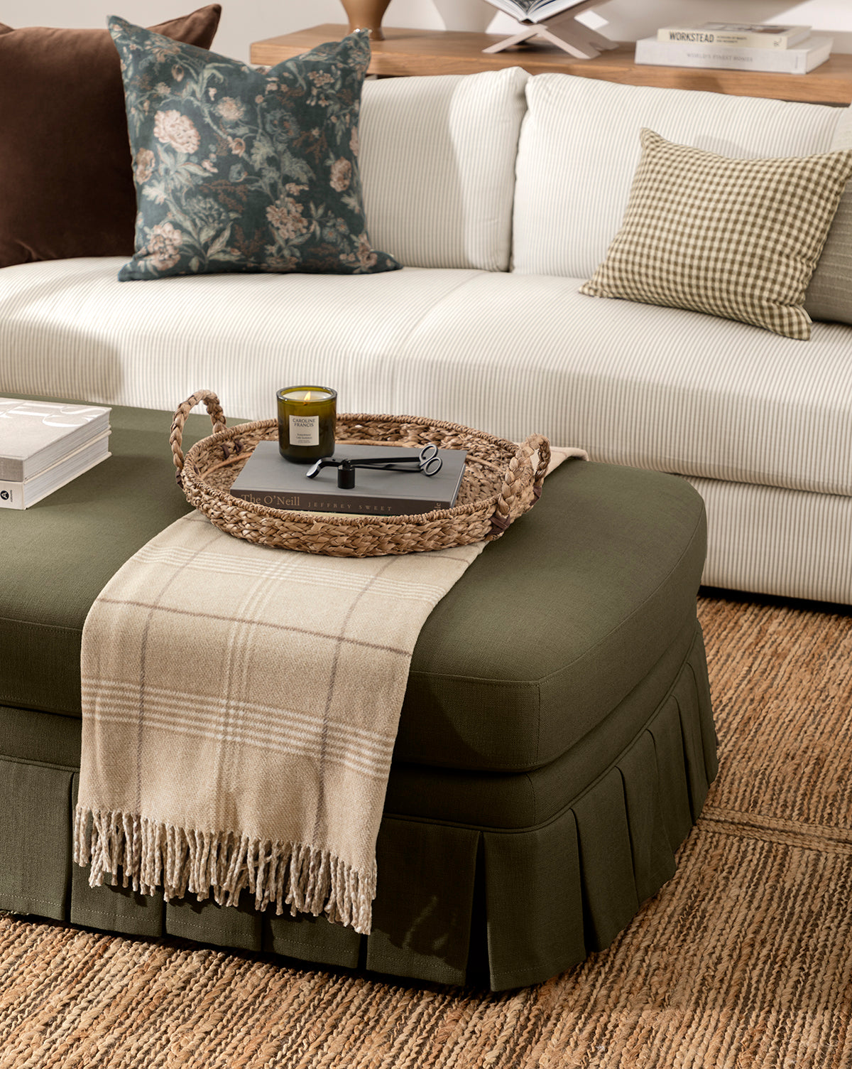 A white sofa with decorative pillows and a green ottoman draped with the Nottingham Throw, plus a woven tray holding a candle, glasses, and books on a textured rug—an inviting accent for any cozy living room, by McGee & Co.