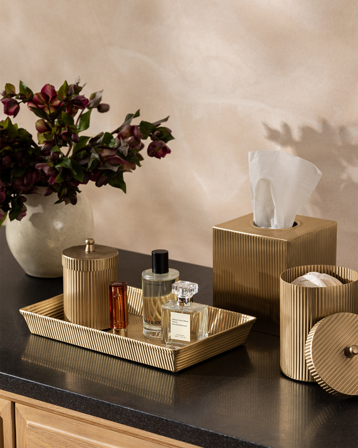 The Calvert Brass Tissue Box Cover sits on a dark countertop with matching gold-toned accessories and a vase of dark red flowers against a beige wall, by McGee & Co.