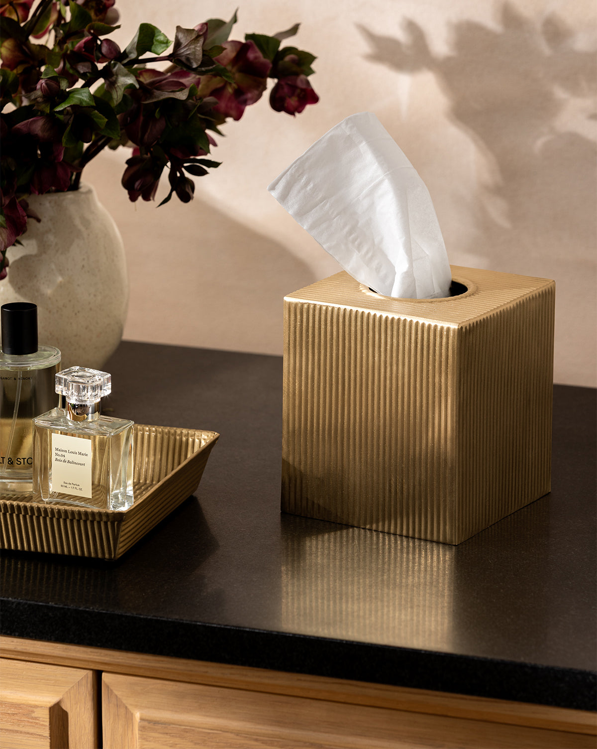 A Calvert Brass Tissue Box Cover sits on a dark countertop beside a tray of perfume bottles, with a vase of deep red flowers visible in the background, by McGee & Co.