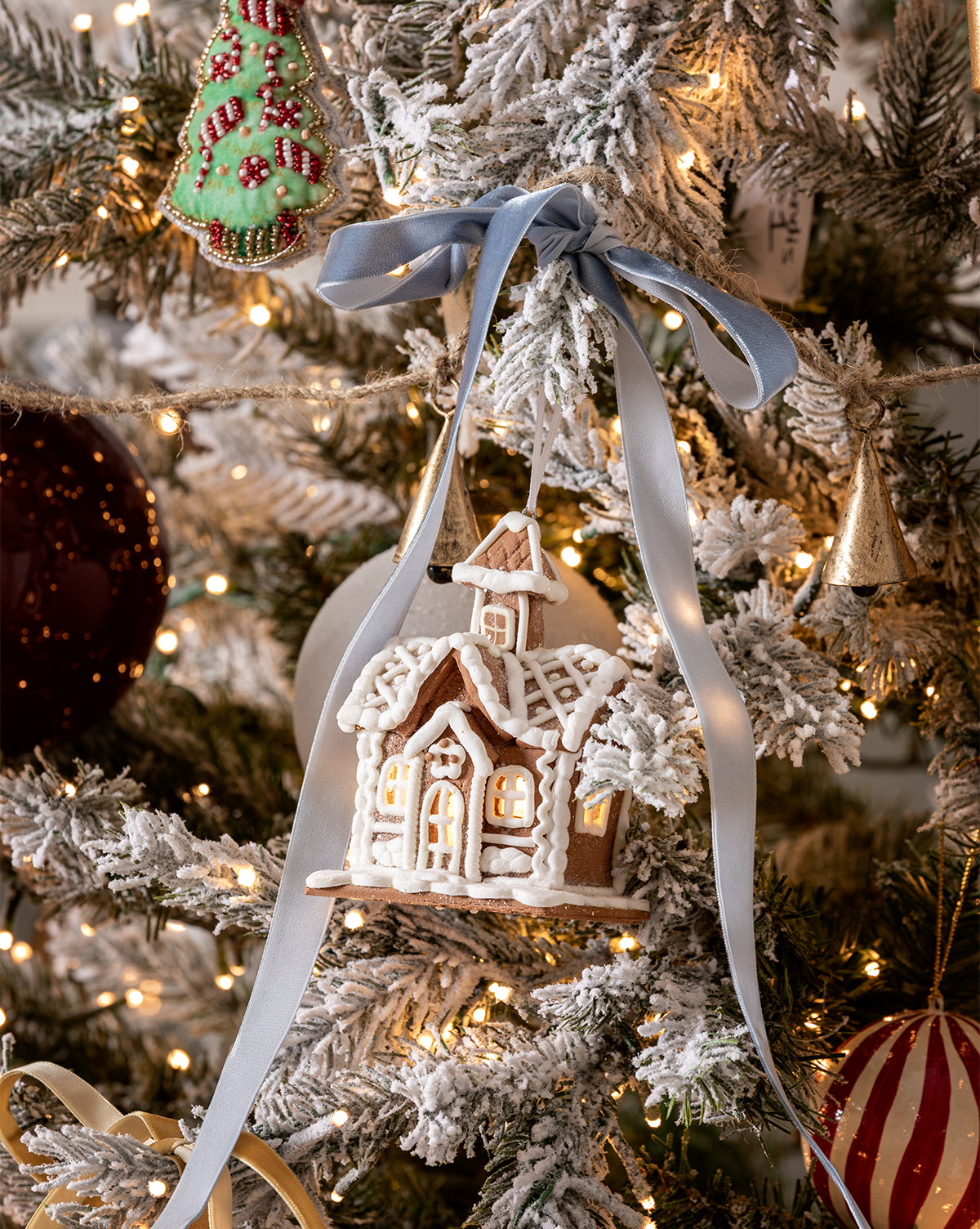 A snowy gingerbread house ornament with white icing and a blue ribbon hangs on the Magical Wonderland Tree, surrounded by lights, a green cookie ornament, and festive holiday decorations, by McGee & Co.