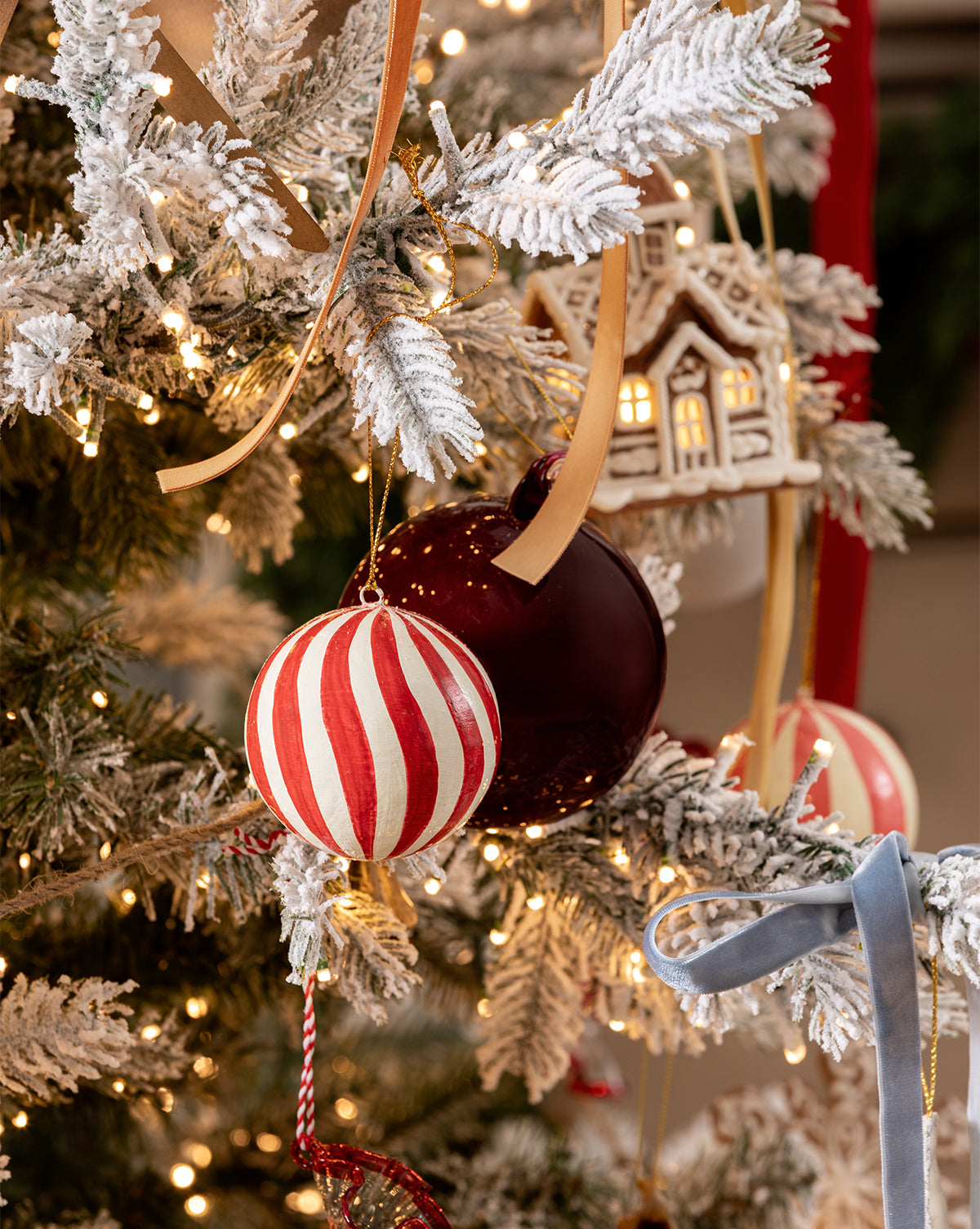 A snow-frosted Christmas tree adorned with red and white striped ornaments, a maroon ball, gold ribbons, warm white string lights, a small house ornament, and 3" Hand-Painted Paper Mache Baubles with Tassel (Set of 3), by McGee & Co.