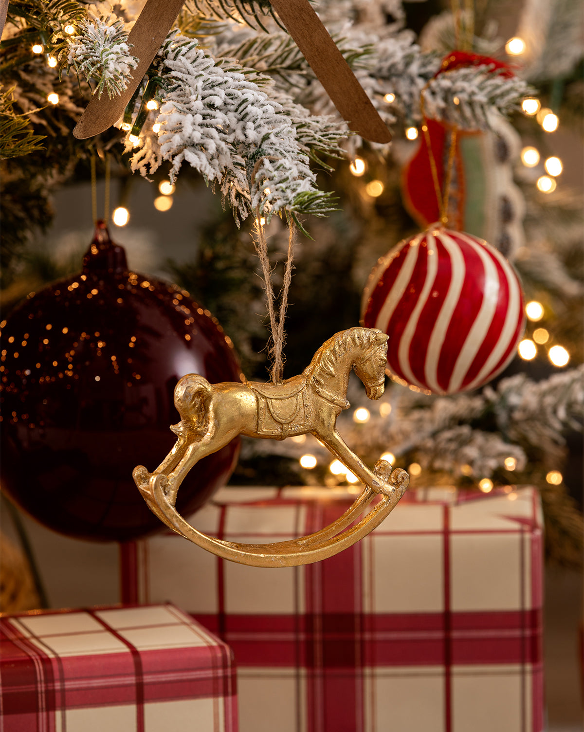 The Gold Rocking Horse Ornament is displayed on a Christmas tree with snowy branches, red and white baubles, and warm lights. Plaid-wrapped presents beneath the tree enhance the festive holiday décor, by McGee & Co.