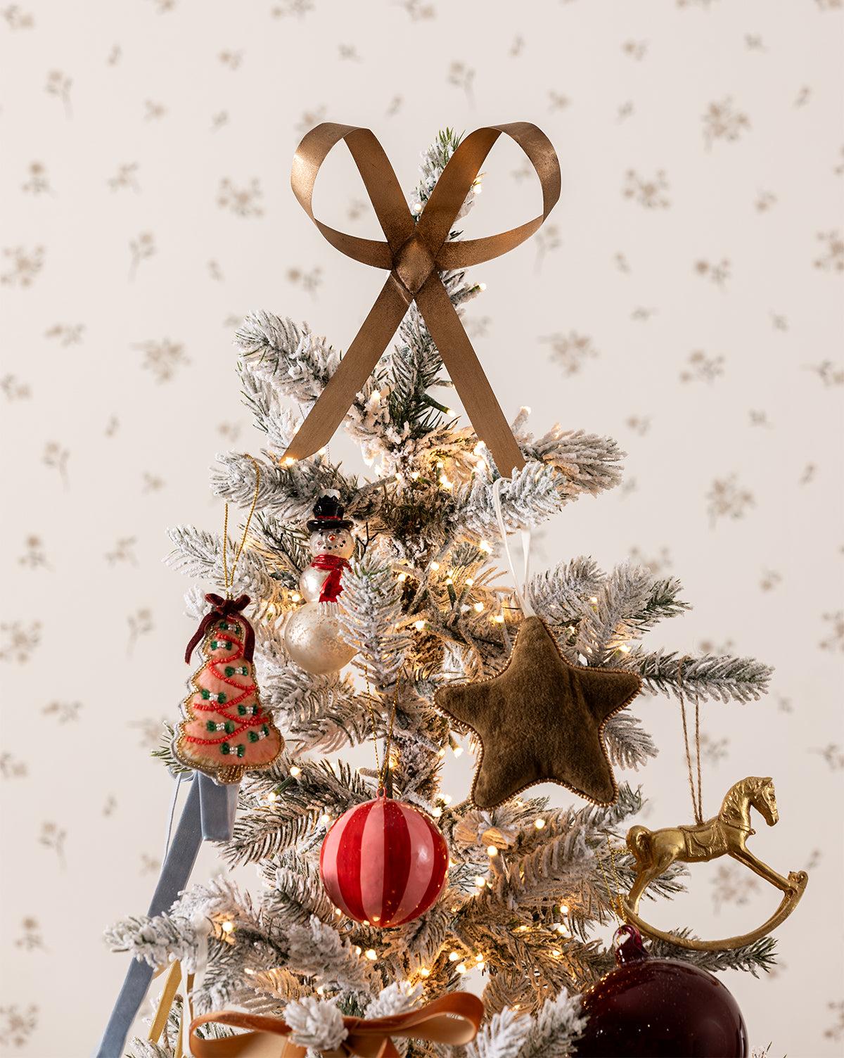 A frosted Christmas tree top decorated with a gold ribbon bow, felt star, snowman, vintage ornament, red-and-white striped ball, glowing white lights, and the Gold Rocking Horse Ornament by McGee & Co.