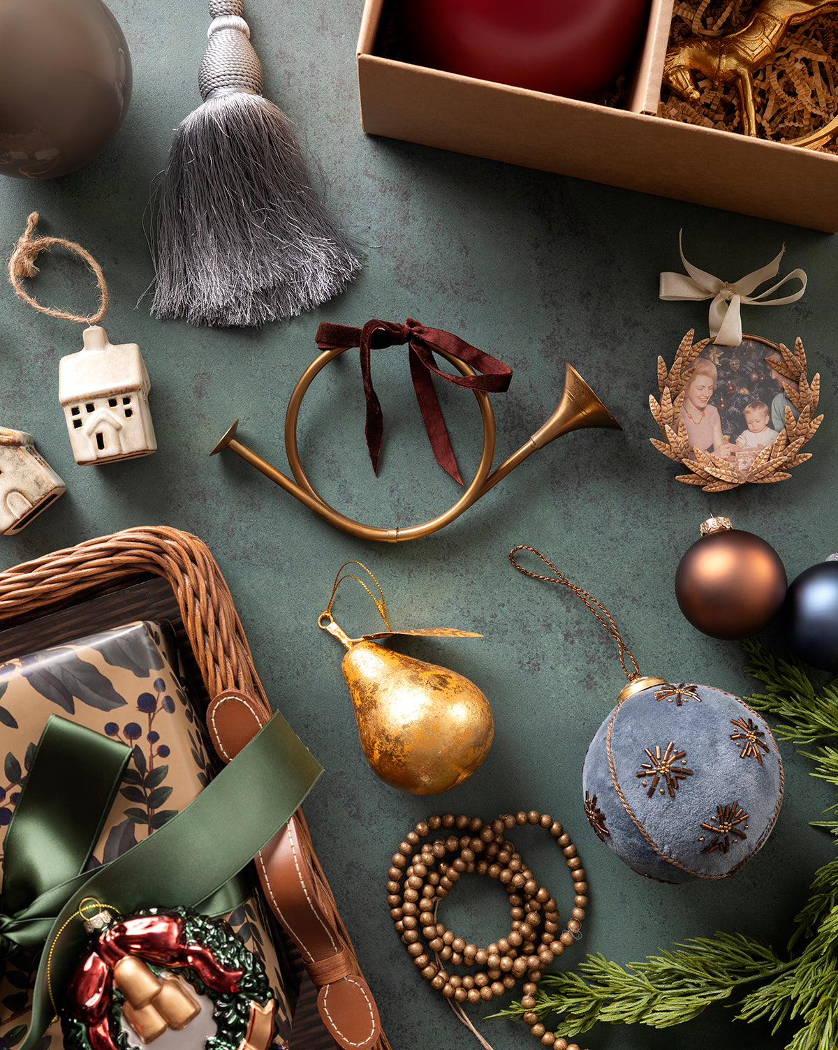 A flat lay of Christmas ornaments, including the Gilded Pear Ornament, beaded garland, brass horn, vintage ornament, baubles, framed photo, ceramic house, tassel, wrapped gifts, and greenery on a green tabletop, by McGee & Co.