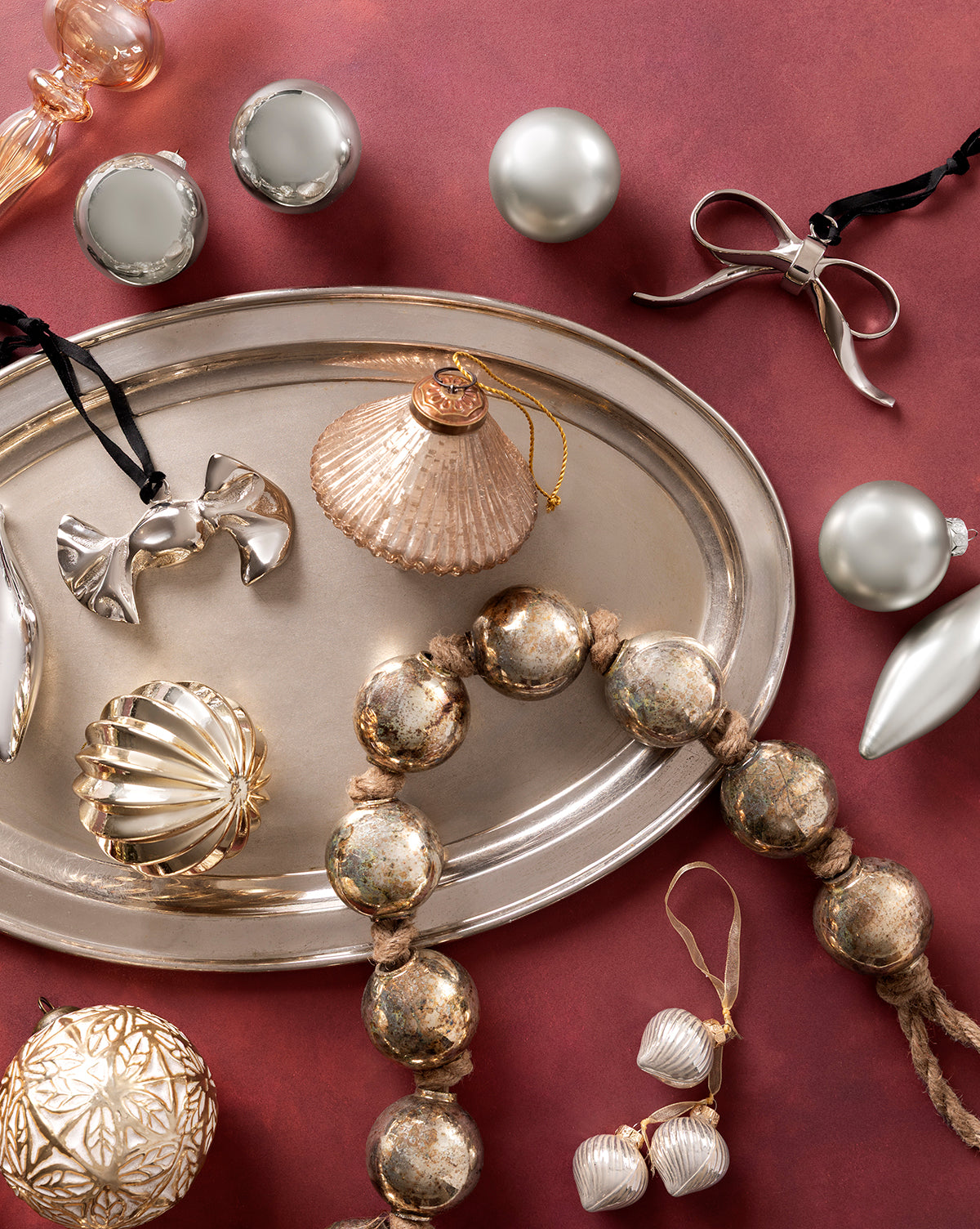 A silver tray with metallic and glass Christmas ornaments—including round, ribbed, and the Silver Bow Ornament—rests on a red surface surrounded by festive décor in silver, gold, and glass, by McGee & Co.