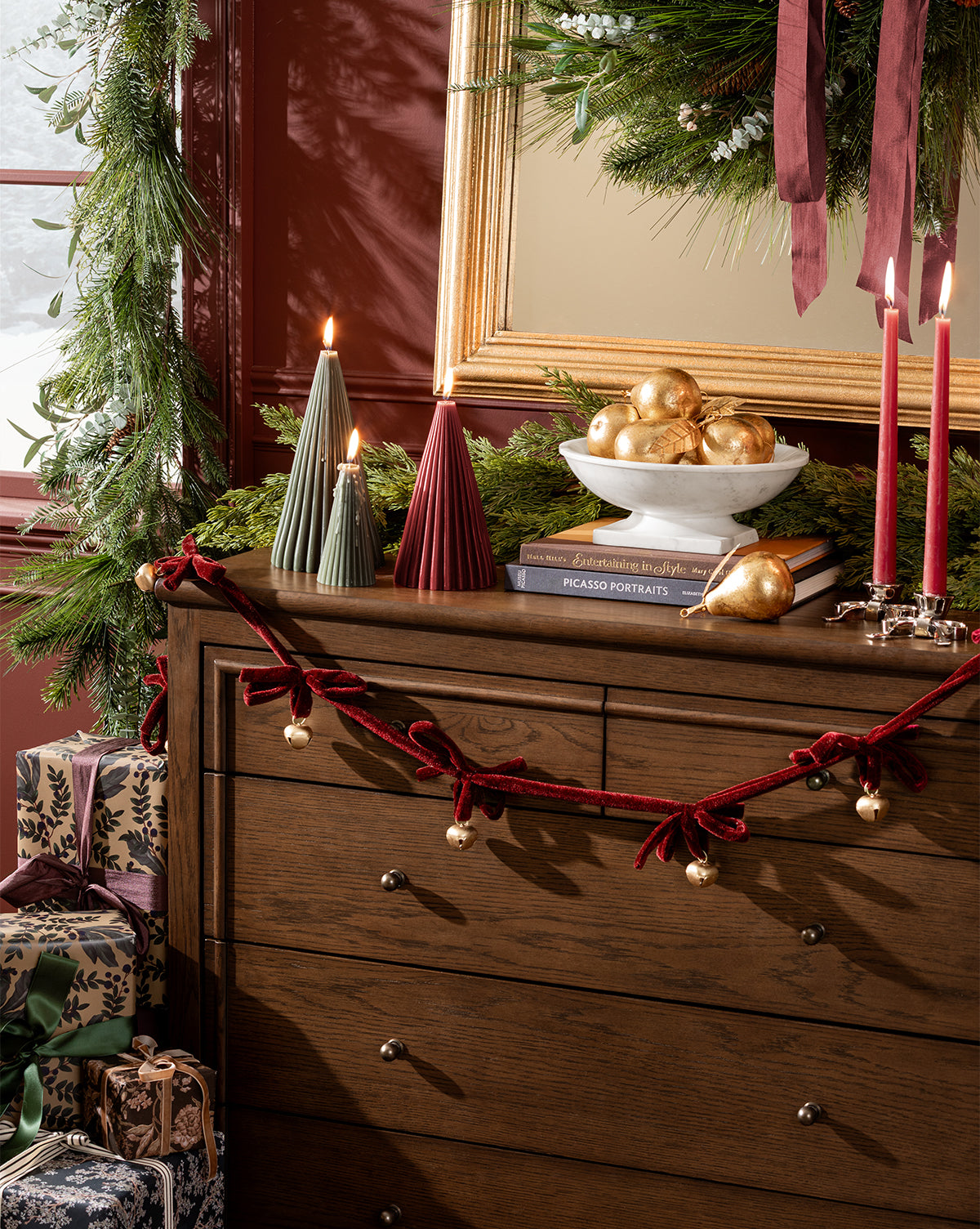 A wooden dresser decorated with the 6' Mini Bows & Bells Garland, candles, wrapped gifts, greenery, gold ornaments, and a wreath creates festive holiday decor featuring red velvet accents and sparkling charm, by McGee & Co.