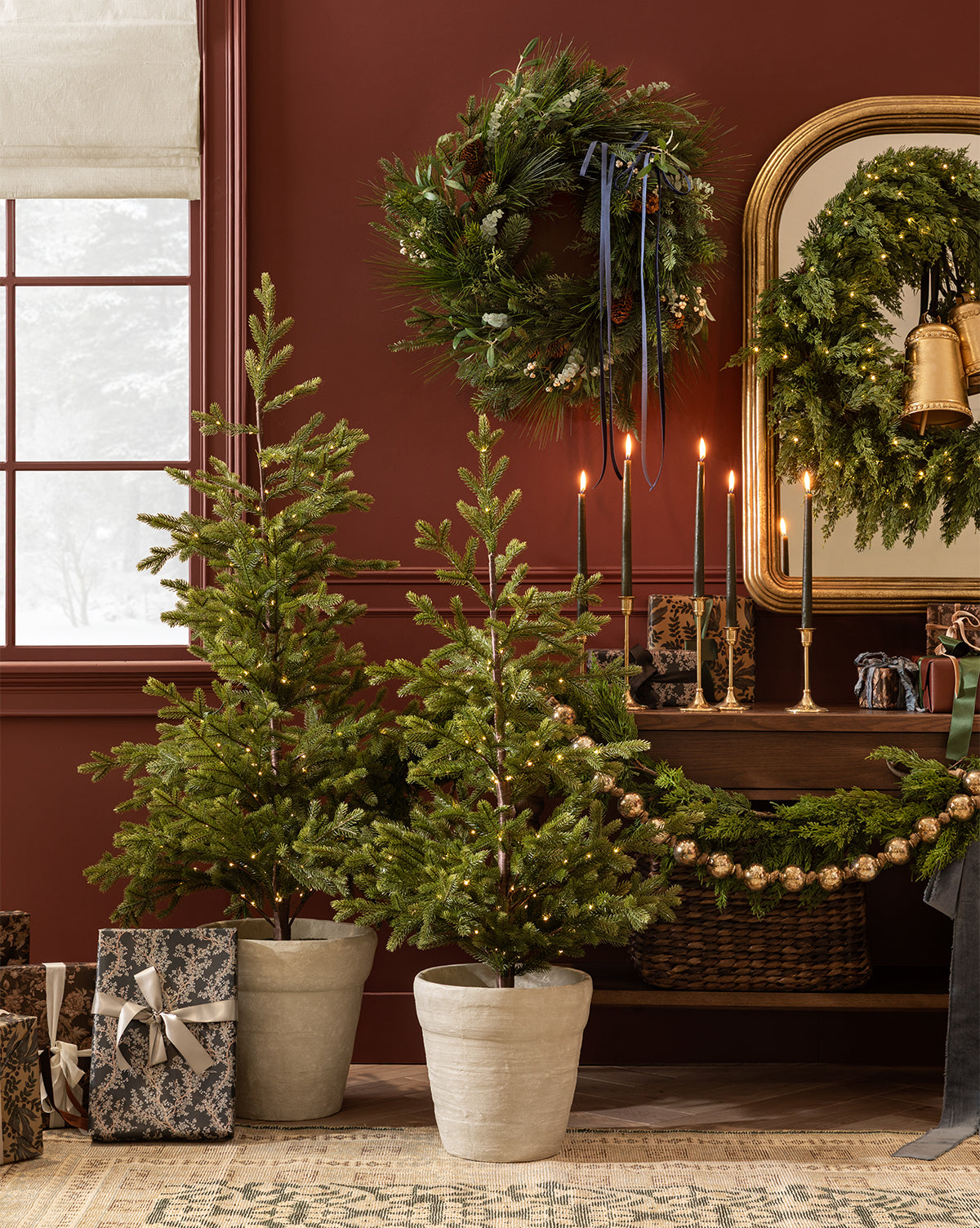 Two Faux Spruce Pre-Lit Potted Trees with string lights stand by wrapped gifts in front of a console table adorned with garland, candles, and greenery, set against a red wall featuring a window and round mirror, by McGee & Co.