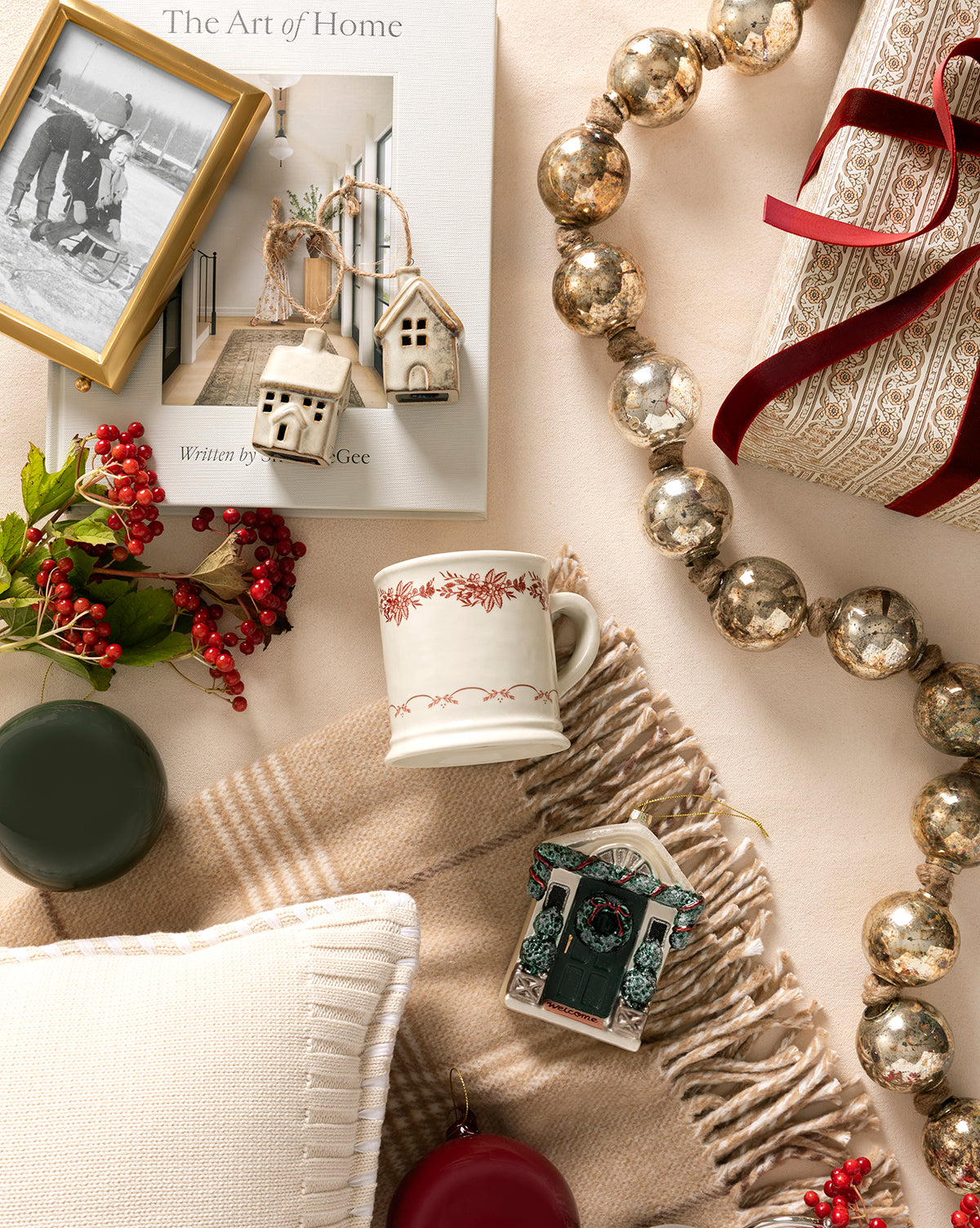 A cozy flat lay with a cream blanket, pillow, festive mug, framed photo, berries, candles, metallic garland, wrapped gift with ribbon, the Glazed Porcelain Houses (Set of 2) as festive accents, and the book “The Art of Home”, by McGee & Co.