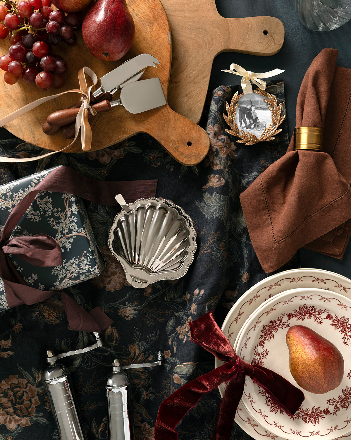 A flat lay of elegant holiday tableware featuring plates with red patterns, a pear, ribbon-tied gifts, brown napkins with gold rings, grapes, the Fromagerie Cheese Knives (Set of 3), and a festive ornament on a dark floral tablecloth, by McGee & Co.