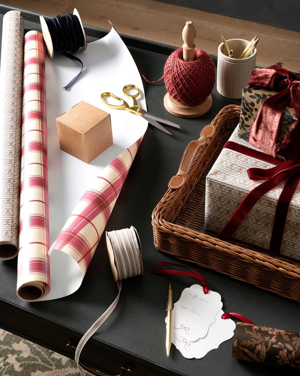 A table with Highland Plaid Gift Wrap, scissors, ribbon, red twine, a cardboard box, a wicker basket of wrapped gifts, pens, and a gift tag, by McGee & Co.