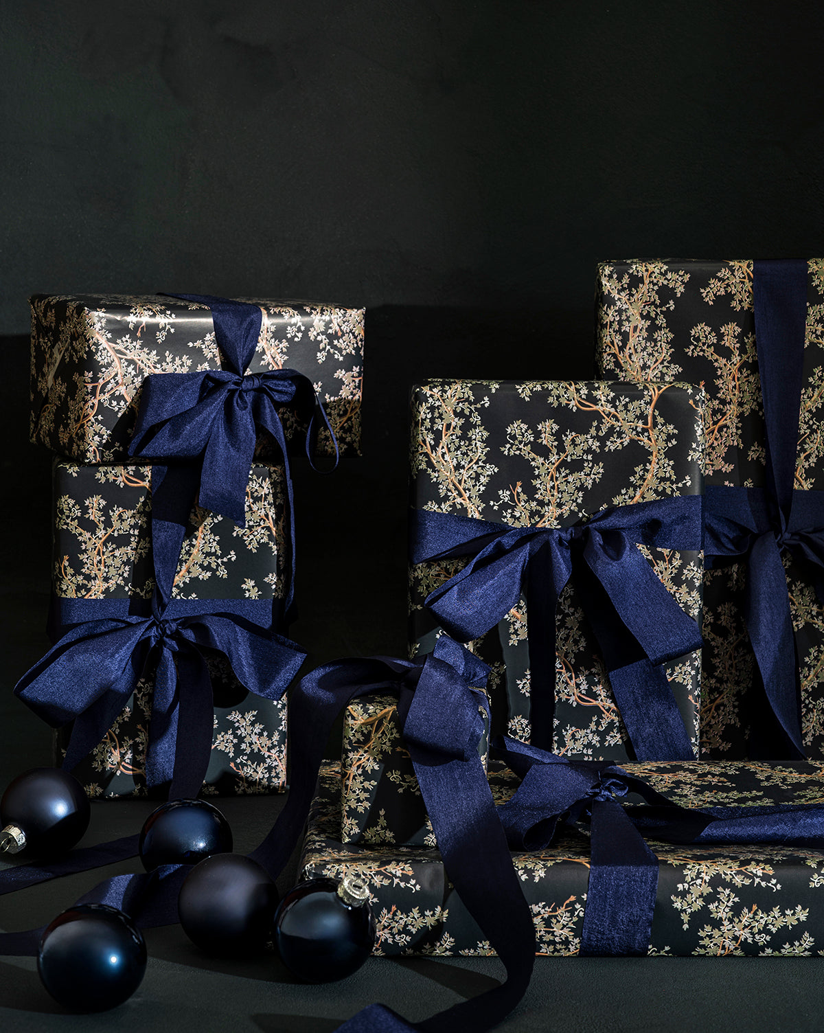 Several gifts wrapped in Antigone Branch Gift Wrap with navy blue ribbons are stacked together, with dark blue ornaments scattered in front, all set against a dark background for an elegant holiday gifting display, by McGee & Co.