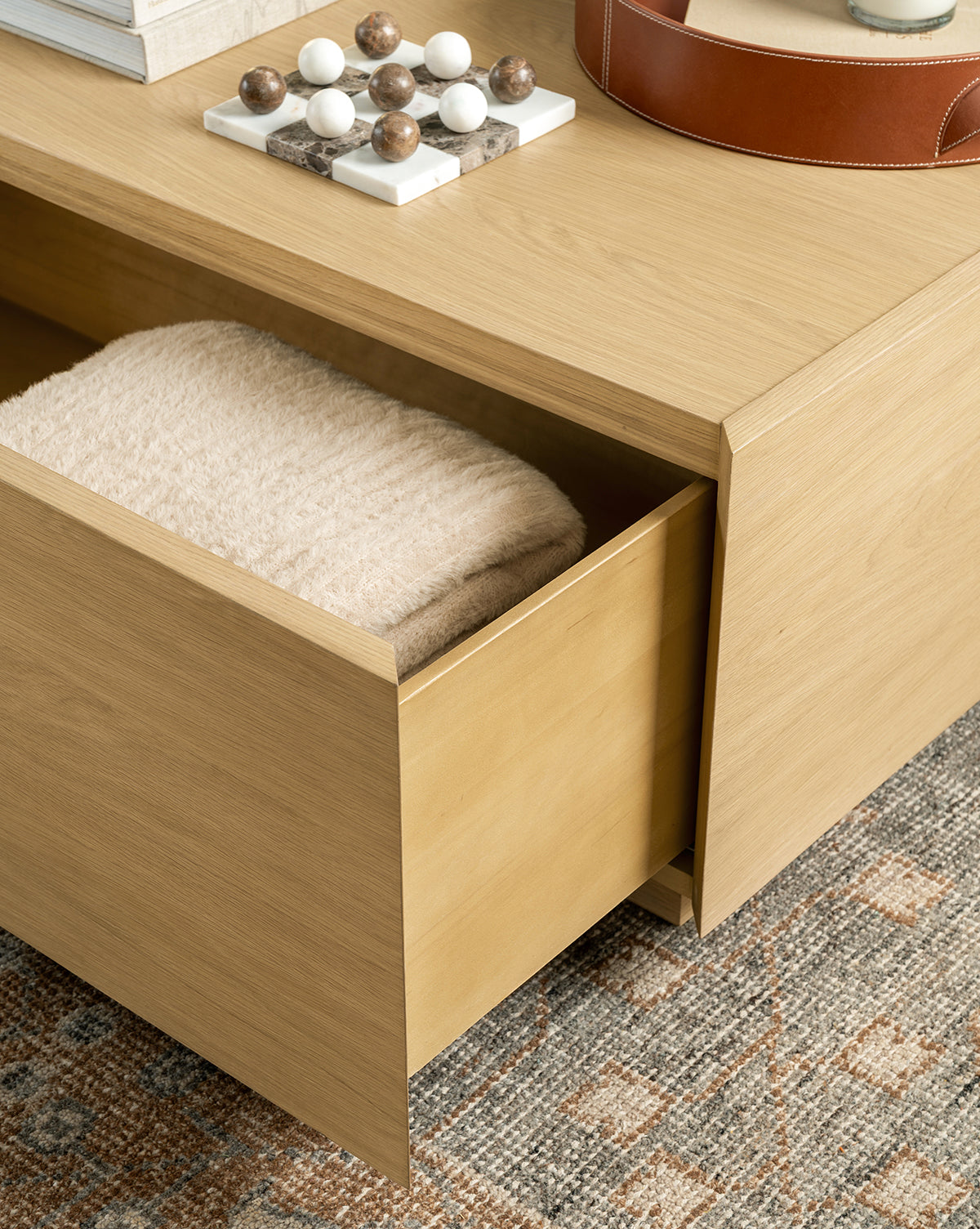 A light wood drawer of the Bentley Coffee Table is partially open, showing a folded beige towel inside. The table sits on a patterned rug with decorative objects and books neatly arranged on top, by McGee & Co.