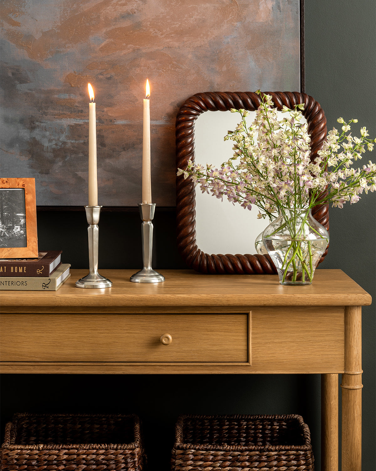 The Hindley Console holds two lit candles in silver holders, a small stack of books, a framed photo, a vase of white flowers, and a braided-frame mirror against a muted wall. Two baskets are placed underneath this entryway console, by McGee & Co.