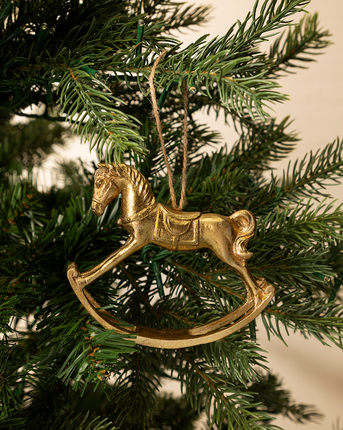 The Gold Rocking Horse Ornament hangs from a string on a green Christmas tree branch, with a softly lit neutral background that adds elegance to your holiday décor, by McGee & Co.