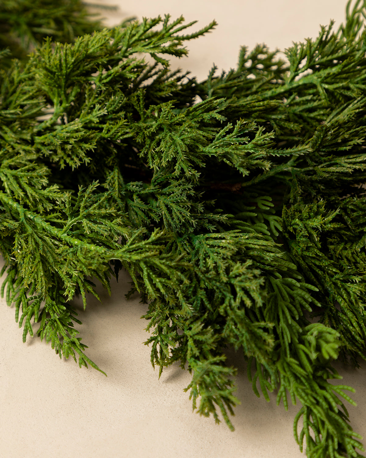 A close-up of the 5' Cedar Faux Garland displays realistic, fresh green cedar branches with fine needle-like leaves arranged on a light beige surface, showcasing an authentic garland effect, by McGee & Co.