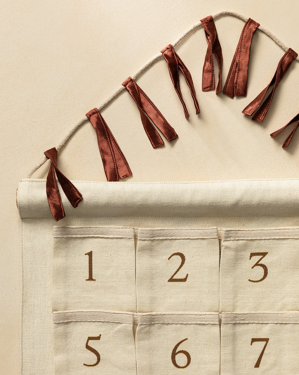 The Advent Calendar is a cotton-linen wall hanging in brown with numbered pockets (1-6 visible), tied to a string with red ribbons. It’s perfect for starting a holiday tradition against a neutral beige background, by McGee & Co.