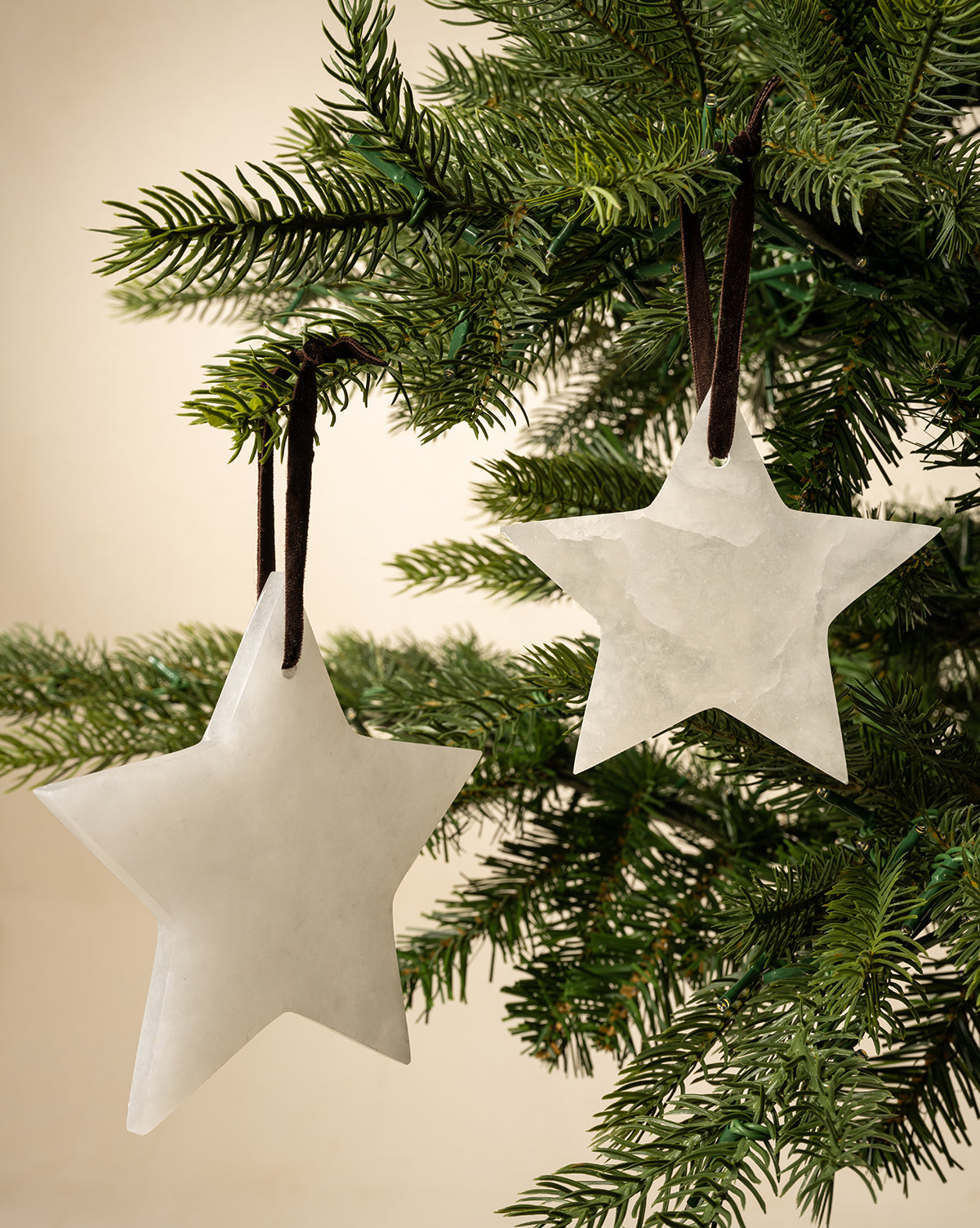 The Alabaster Star Ornaments (Set of 2) hang from brown ribbons on a green Christmas tree, bringing elegant style to your festive décor against a neutral beige background, by McGee & Co.