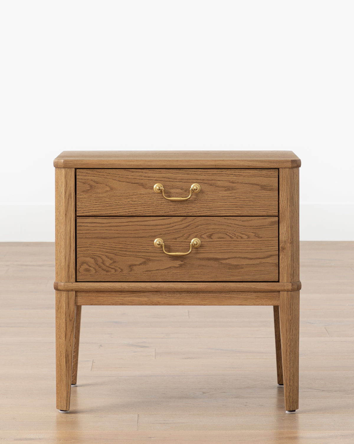 The Alessandra Nightstand by Makerspalm, crafted from solid oak, features two drawers with brass handles and stands on four tapered legs, shown against a light wall and wood floor. McGee & Co.