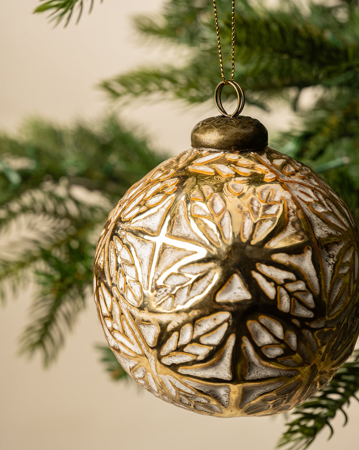 The Alessandro Glass Ornament features a gold leaf and snowflake design, shown in close-up as it hangs from a green Christmas tree branch for festive holiday decor, by McGee & Co.