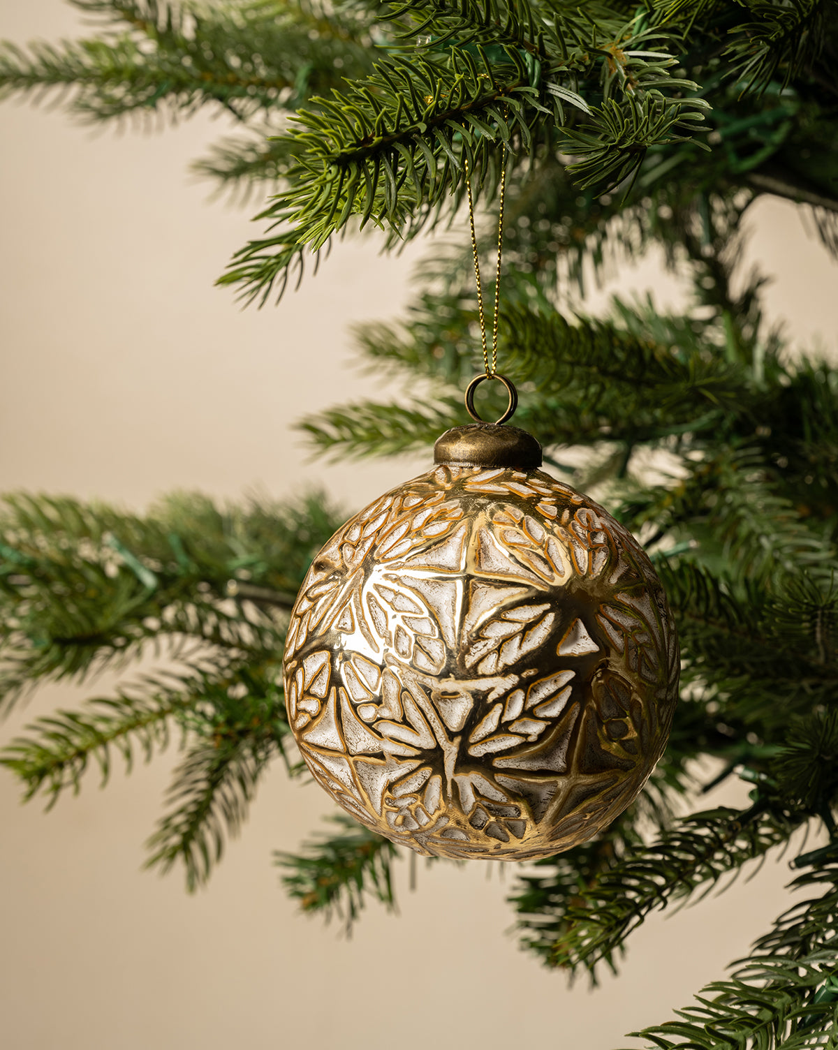 The Alessandro Glass Ornament, featuring an intricate gold pattern, hangs from a green pine branch against a neutral backdrop, bringing timeless charm to your holiday decor, by McGee & Co.