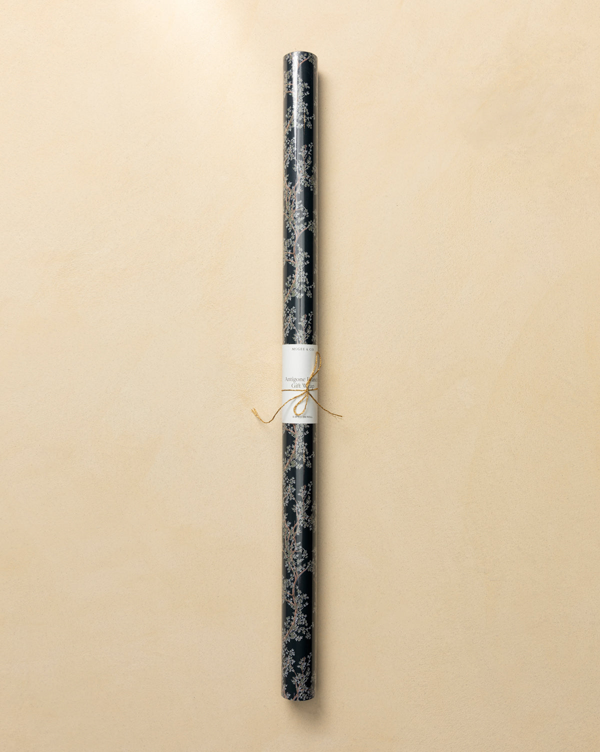 A roll of Antigone Branch Gift Wrap featuring a dark, nature-inspired design with a gold botanical pattern, tied with a thin gold string and labeled, displayed on a light beige background, by McGee & Co.