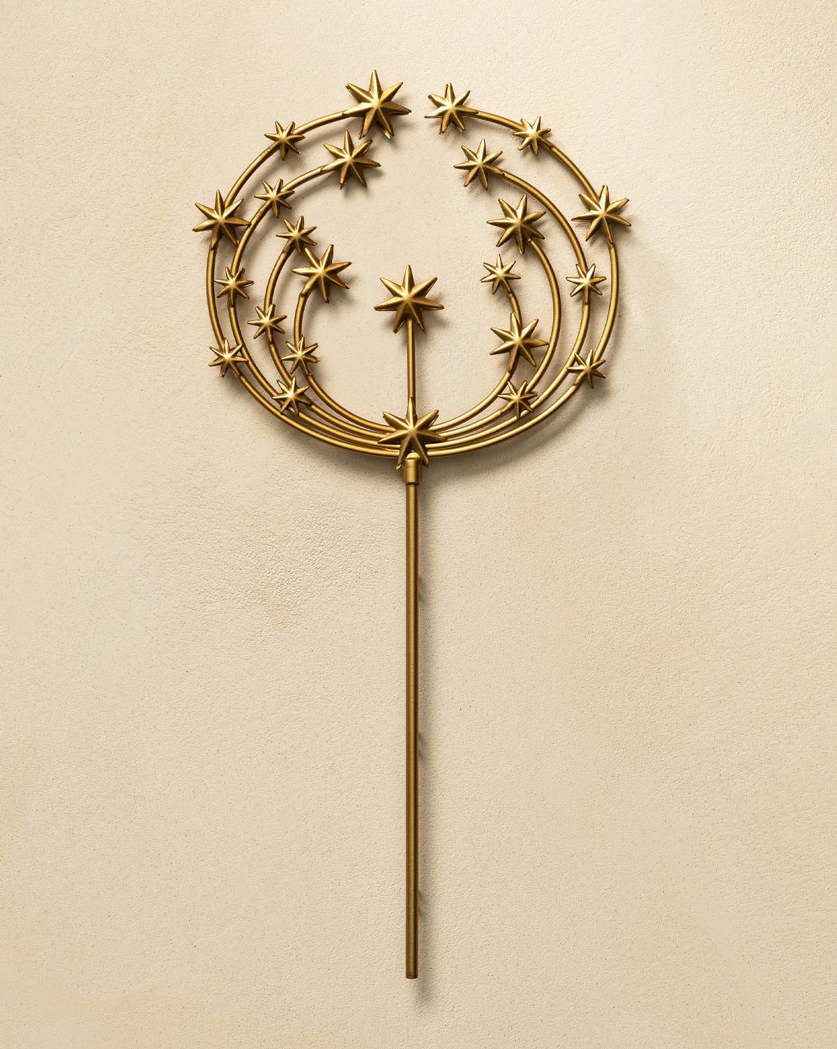 The Astra Brass Tree Topper features a gold metal wand with an antique brass finish, a round top adorned with stars, and three concentric arcs radiating from a central star, set on a beige textured background—ideal for holiday decor, by McGee & Co.