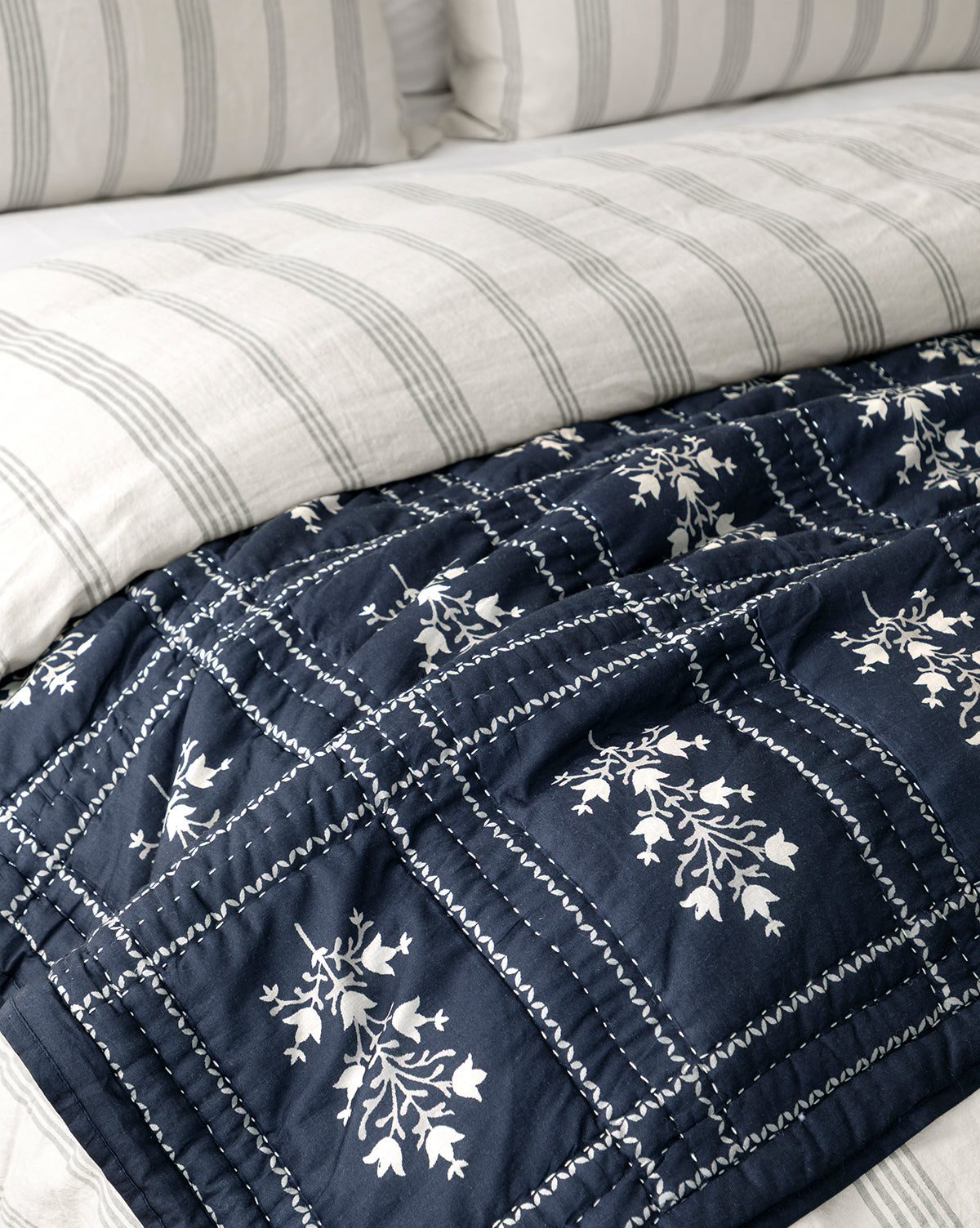 A close-up of a neatly made bed with white and gray striped bedding, topped with the Atherton Quilt—navy blue with white floral patterns, decorative stitching, and handmade artistry, by McGee & Co.