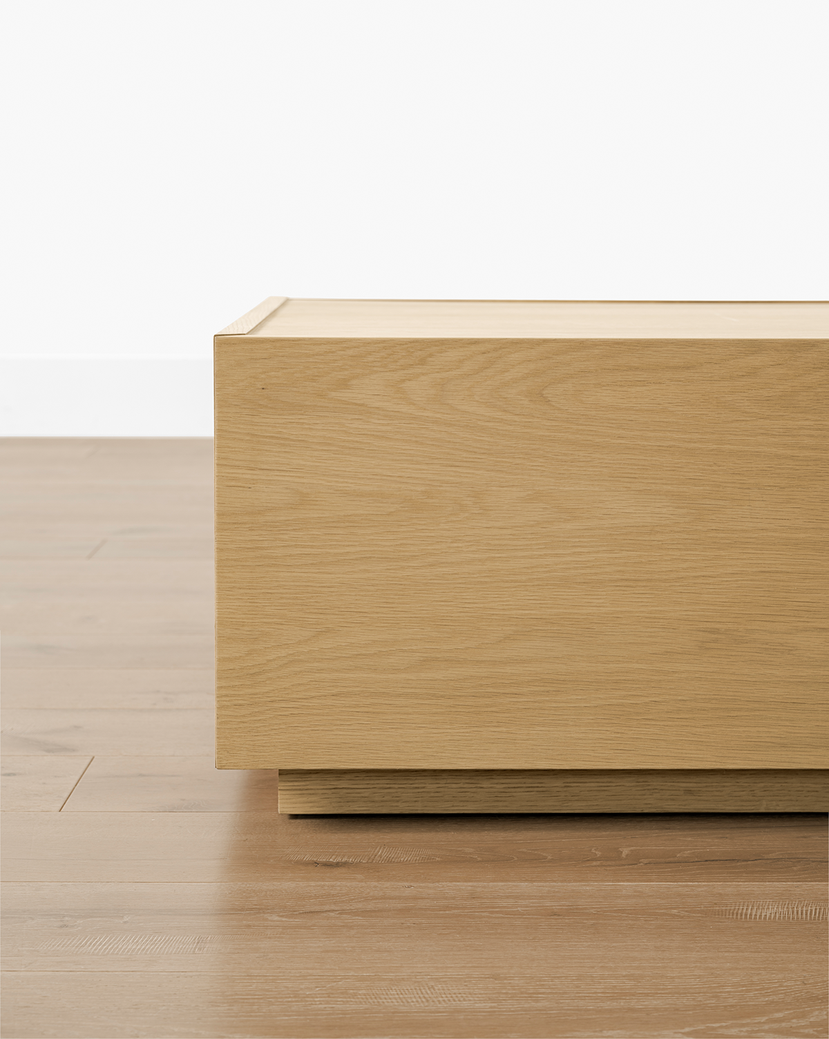 A close-up of the Bentley Coffee Table, featuring a rectangular, light oak finish. Positioned on a wooden floor by a white wall, only its stylish corner is visible, by McGee & Co.