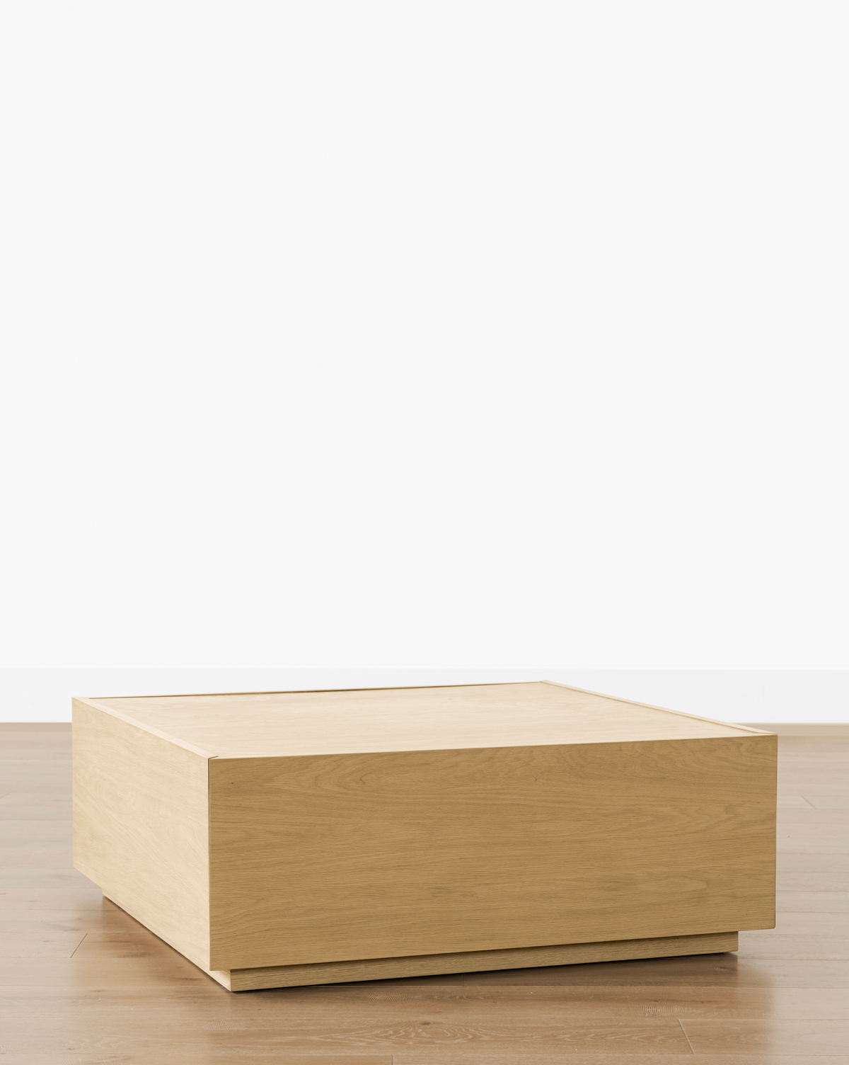 The Bentley Coffee Table is a minimalist, square-shaped design in light oak, featuring clean lines and a solid, box-like look. Perfect for modern spaces, it sits beautifully on wooden floors against plain white walls, by McGee & Co.