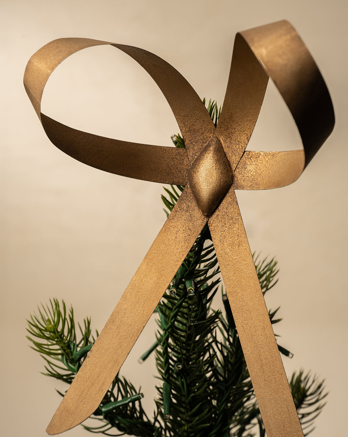 The Brass Bow Tree Topper, featuring an antique brass finish, sits in a classic bow atop the green tip of a Christmas tree against a neutral background, by McGee & Co.