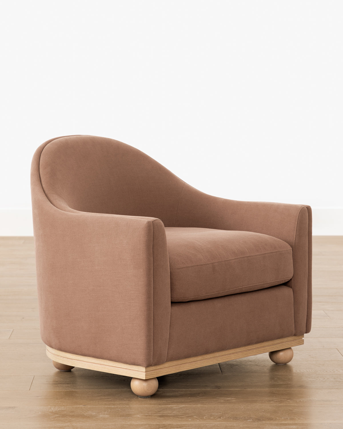The Bunnell Lounge Chair (Ready to Ship) features a curved back, armrests, and light brown upholstery. Its modern traditional design sits on a wooden base with round feet, displayed on a light wood floor against a plain white wall by McGee & Co.