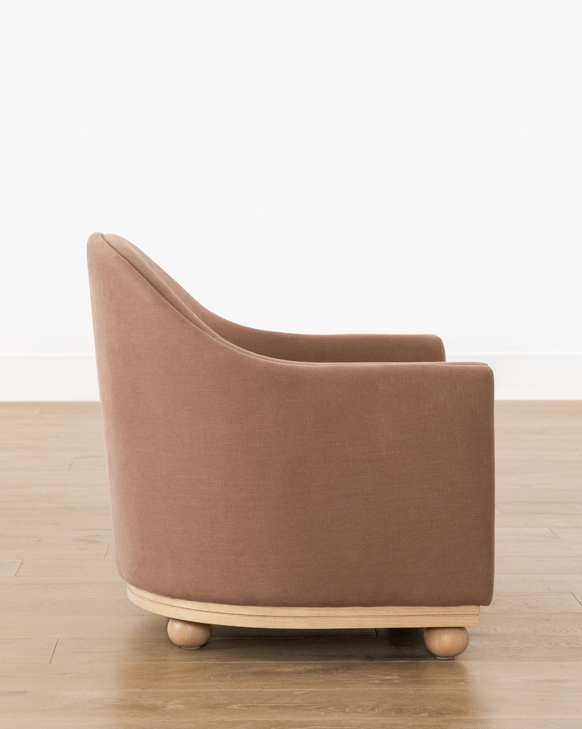 The Bunnell Lounge Chair (Ready to Ship) features modern brown upholstery, a rounded back and arms, and a light wood base with ball-shaped feet. Its shown on a wooden floor against a white wall by McGee & Co.