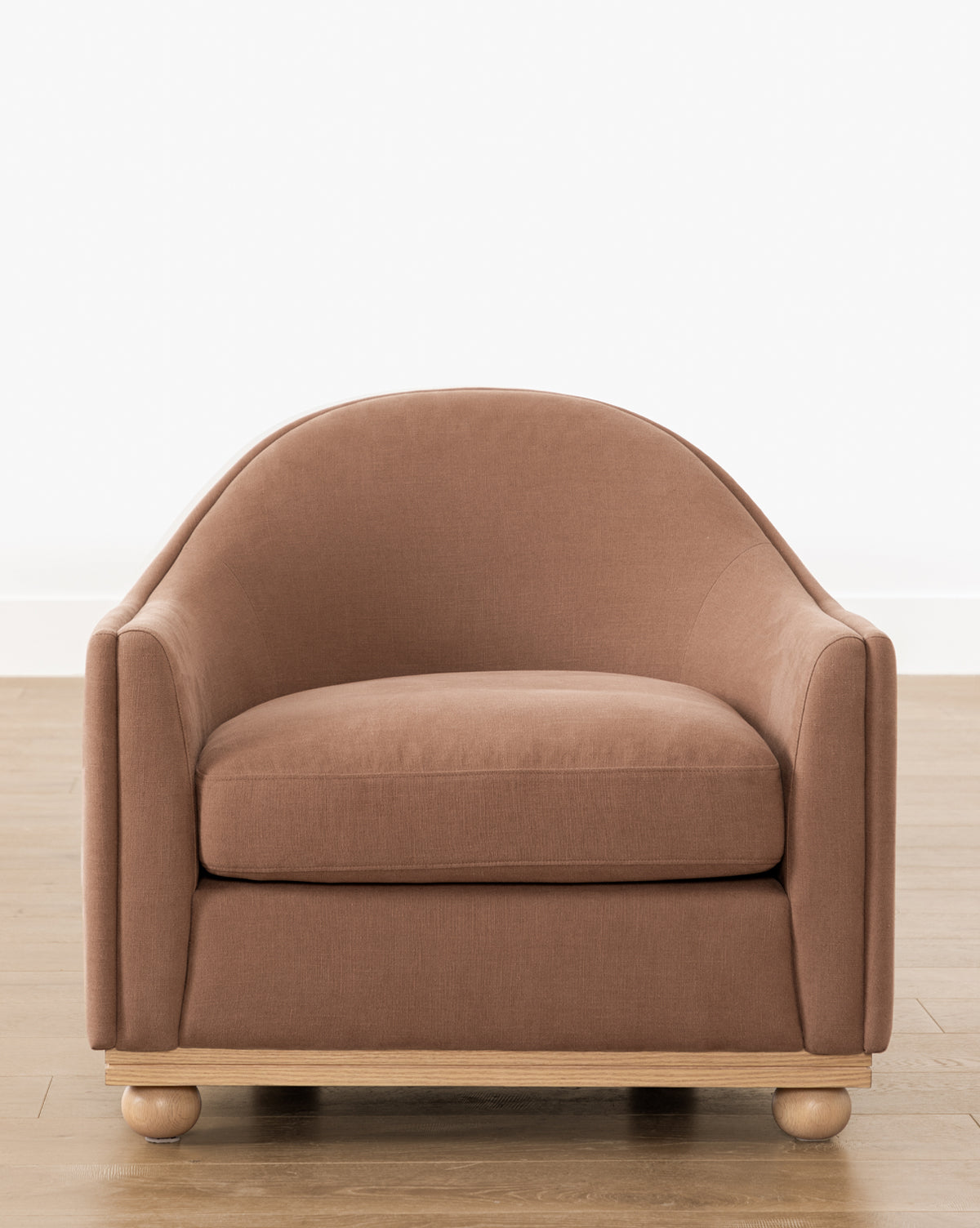 The Bunnell Lounge Chair (Ready to Ship) by Makerspalm features brown upholstery, a rounded backrest, low wide seat, and light wooden base with round feet, shown on a wooden floor against a white wall. McGee & Co.